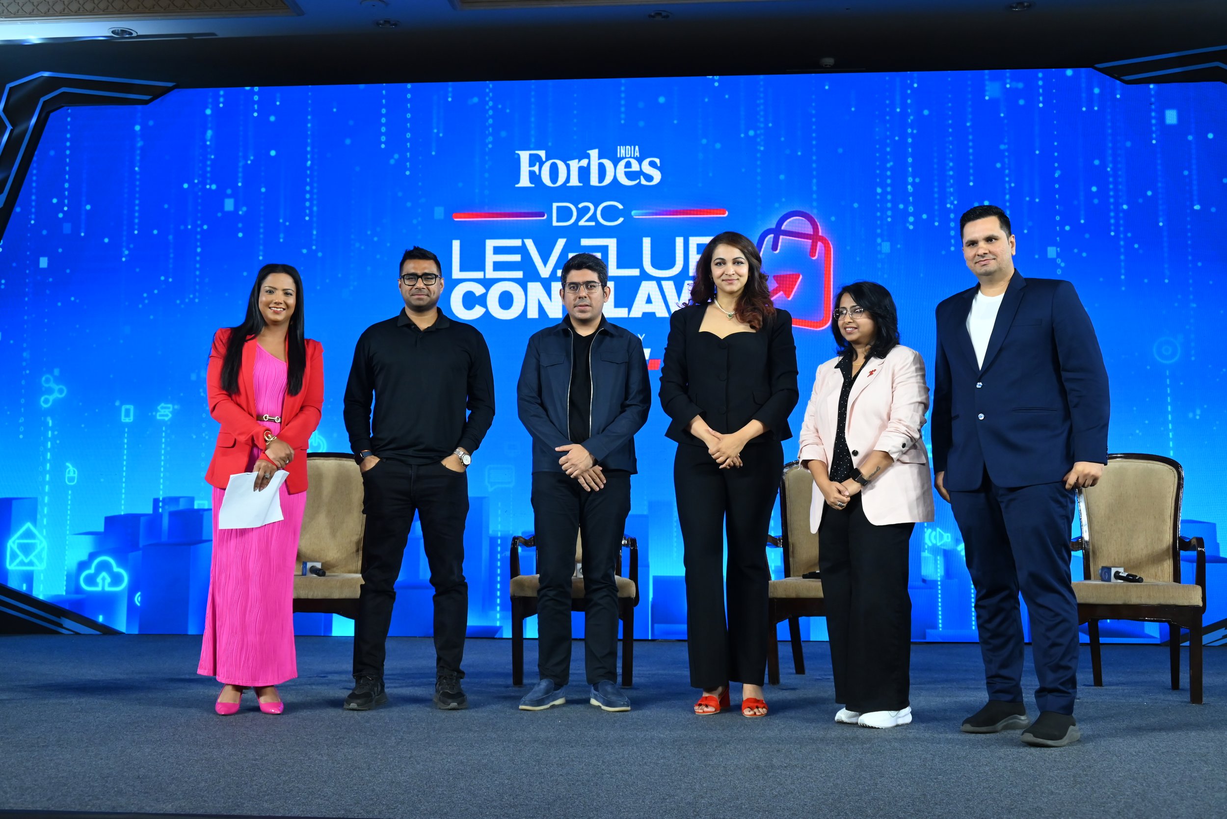 A group of six people standing on stage in front of a blue digital-themed backdrop that reads "Forbes India D2C Level Up Conference." The group is dressed in business casual to formal attire, with three women and three men.