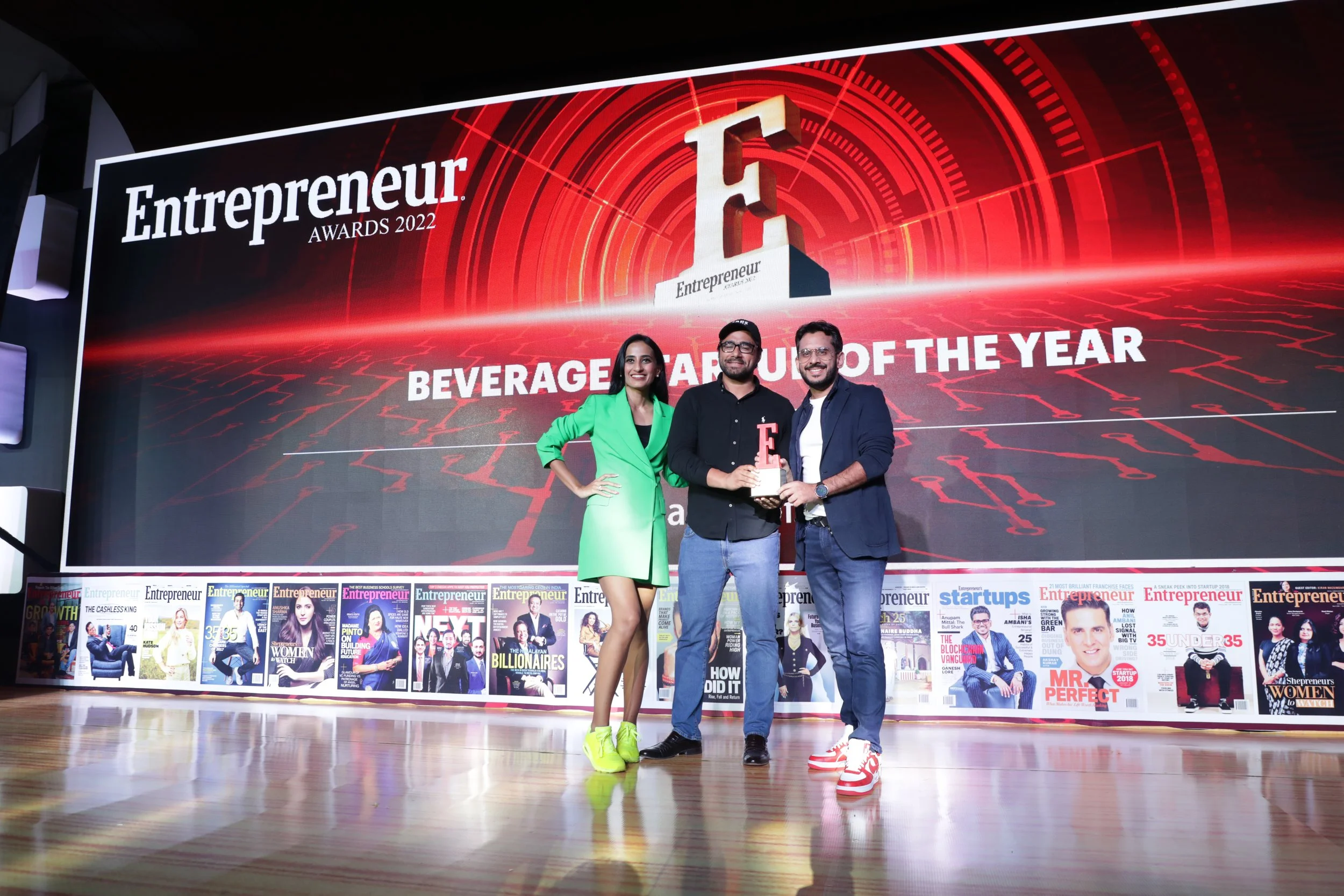 Three people on stage at the Entrepreneur Awards 2022, holding a trophy for Beverage Brand of the Year. The woman wears a green blazer dress and neon yellow shoes, the man in the middle wears a black shirt and jeans, and the man on the right wears a 