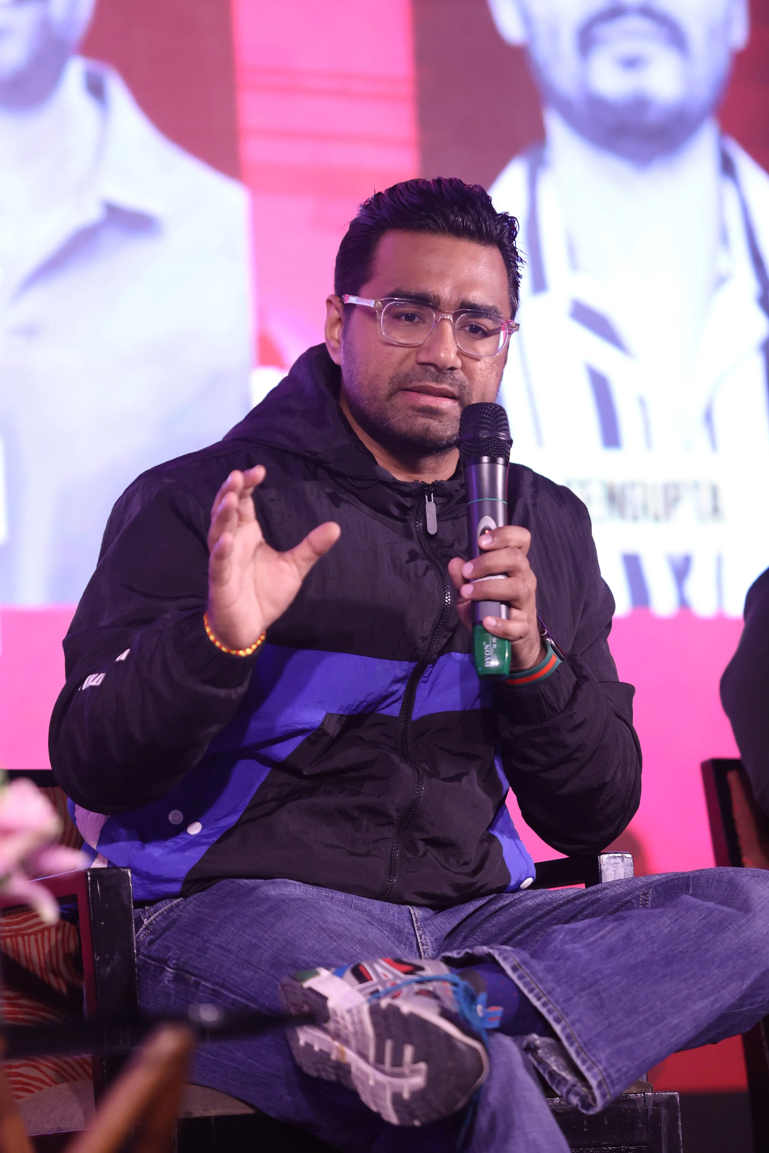 A man wearing glasses, a black jacket with blue accents, and jeans, sitting on a chair and speaking into a microphone during a panel or event, with a large screen in the background showing part of his image.
