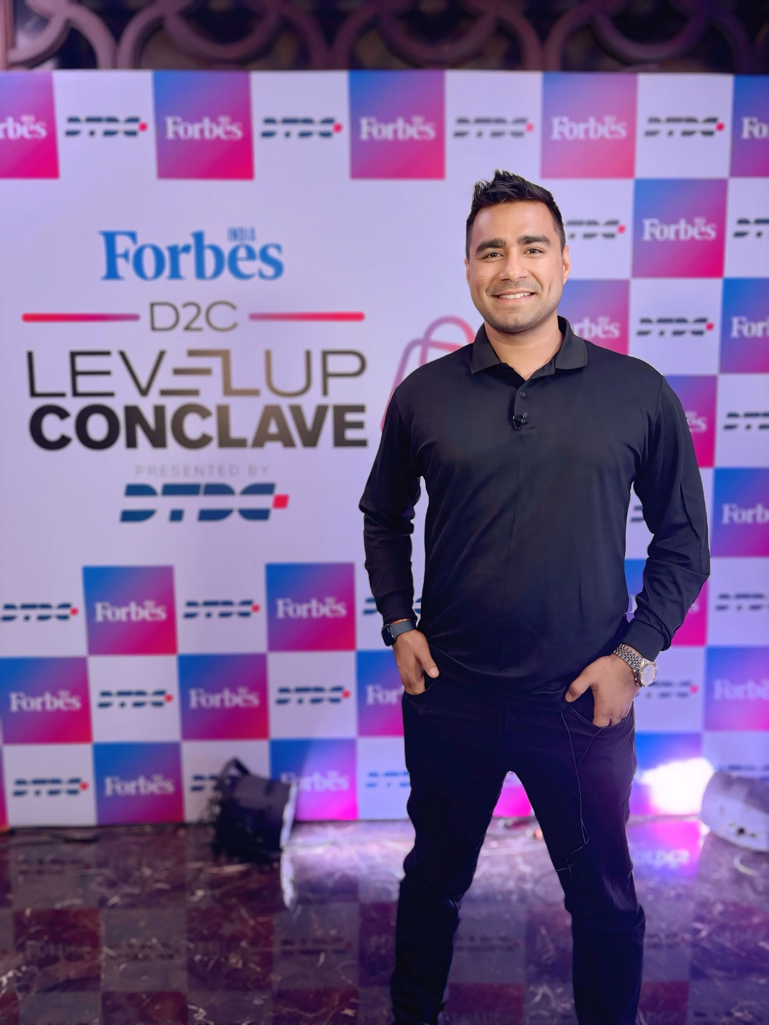 A man in a black long-sleeve shirt and black pants standing in front of a backdrop with multiple Forbes logos and a sign that says "Level Up Conclave".