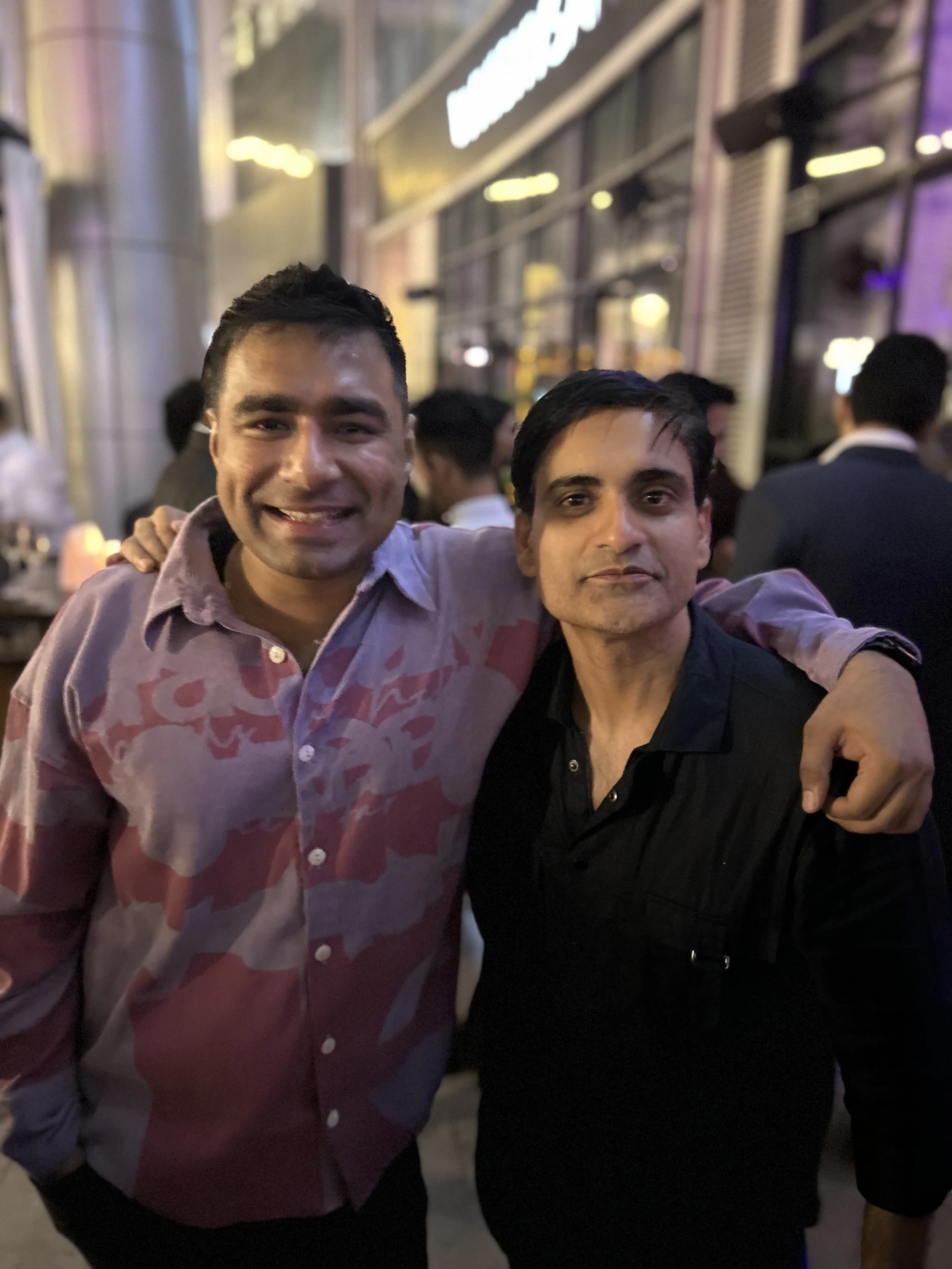 Two men smiling and posing with their arms around each other at a nighttime outdoor event or party, with a crowded background and city lights.