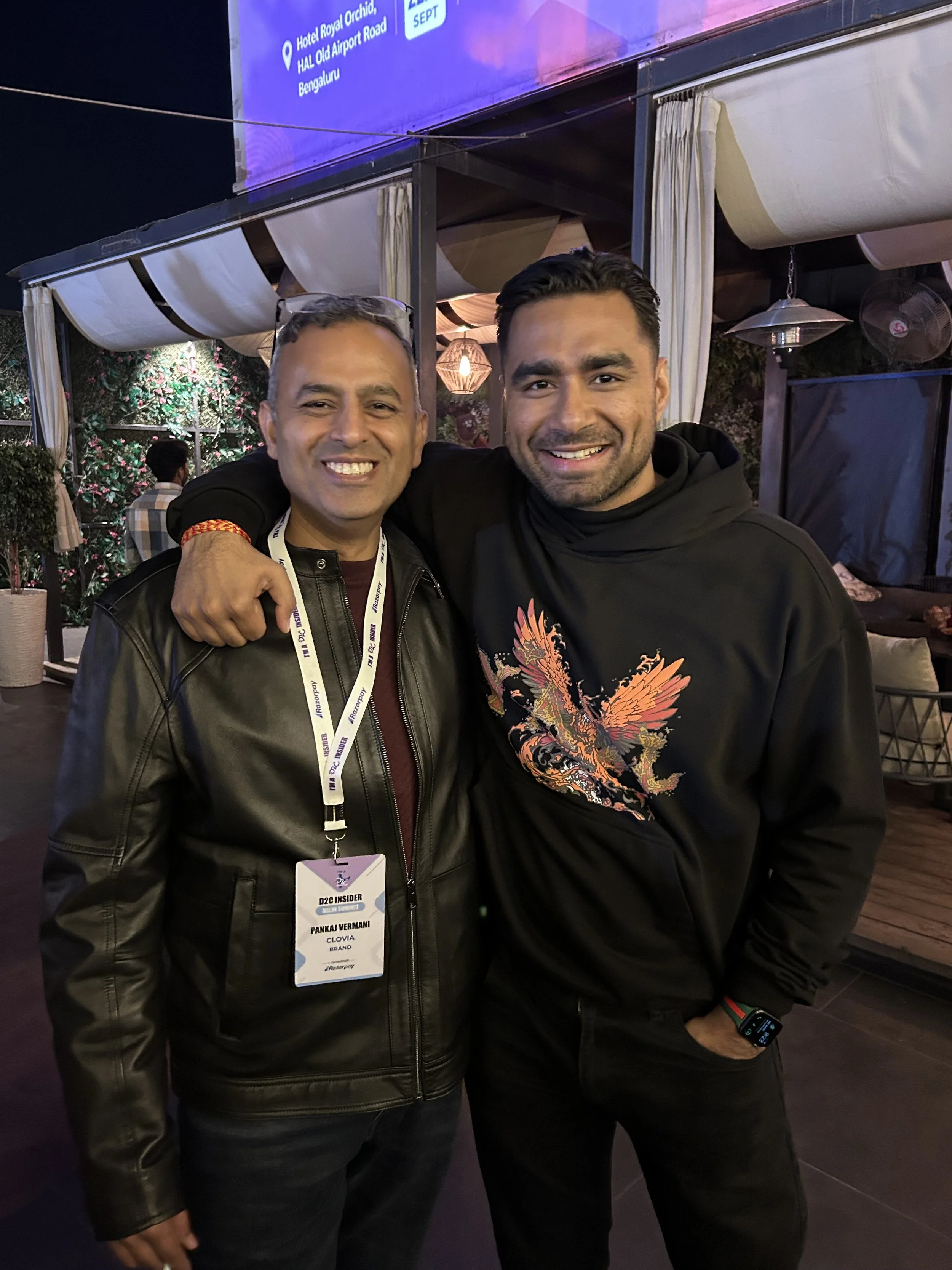 Two men smiling and posing together at an evening event, with one man wearing a black leather jacket and the other in a black hoodie with a colorful dragon design, at a venue with draped curtains and floral decorations in the background.