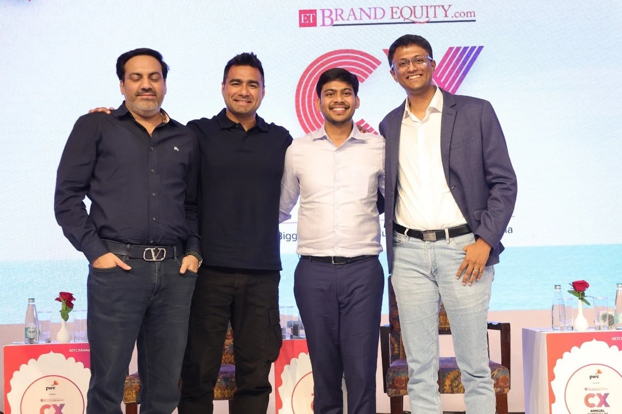 Four men standing together on stage at a corporate event, smiling for a photo. They are in front of a backdrop with the text 'ET Brand Equity' and a logo, and there are tables with water bottles and flowers nearby.