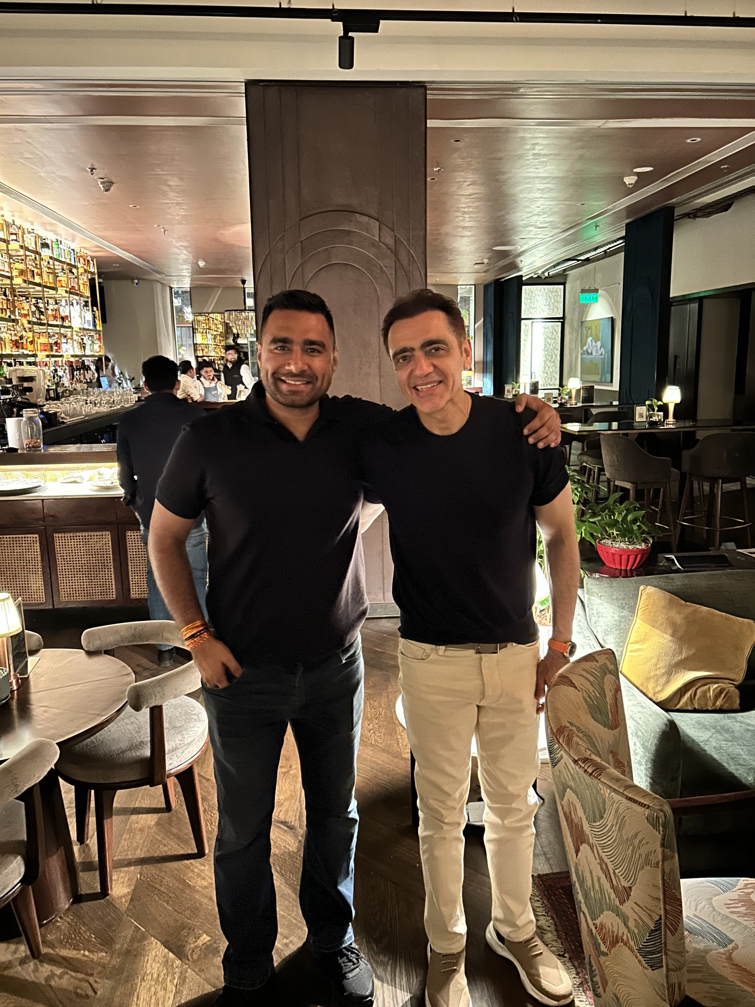 Two men standing side by side in a restaurant, smiling at the camera with their arms around each other's shoulders.