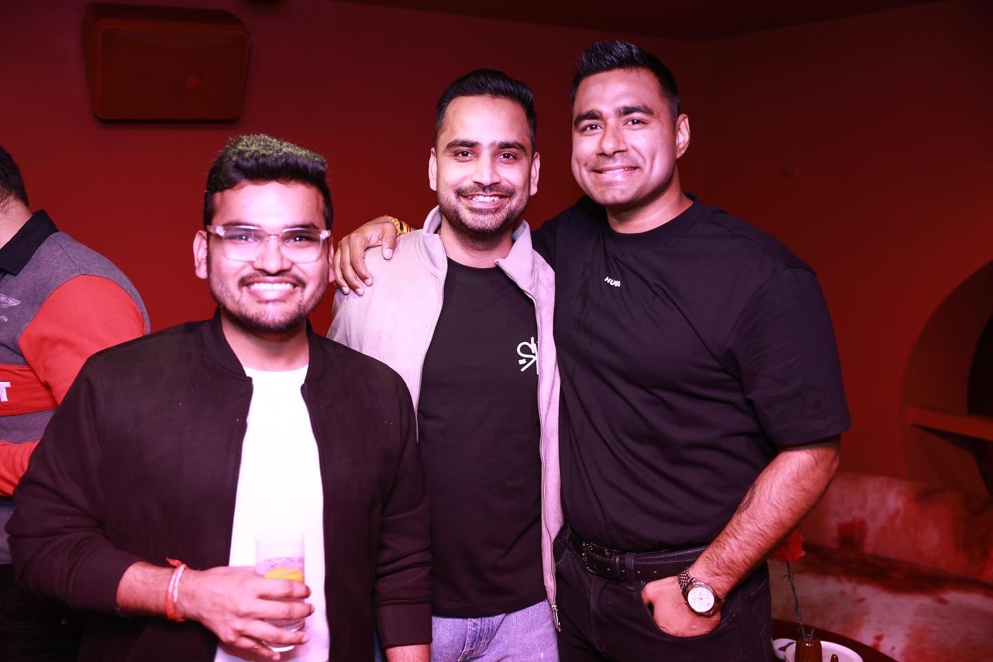 Three men smiling, standing close together at a social event. The man on the left is wearing glasses, a black jacket, and holding a glass of beverage. The man in the middle has a beard, is wearing a light-colored jacket over a black shirt. The man on