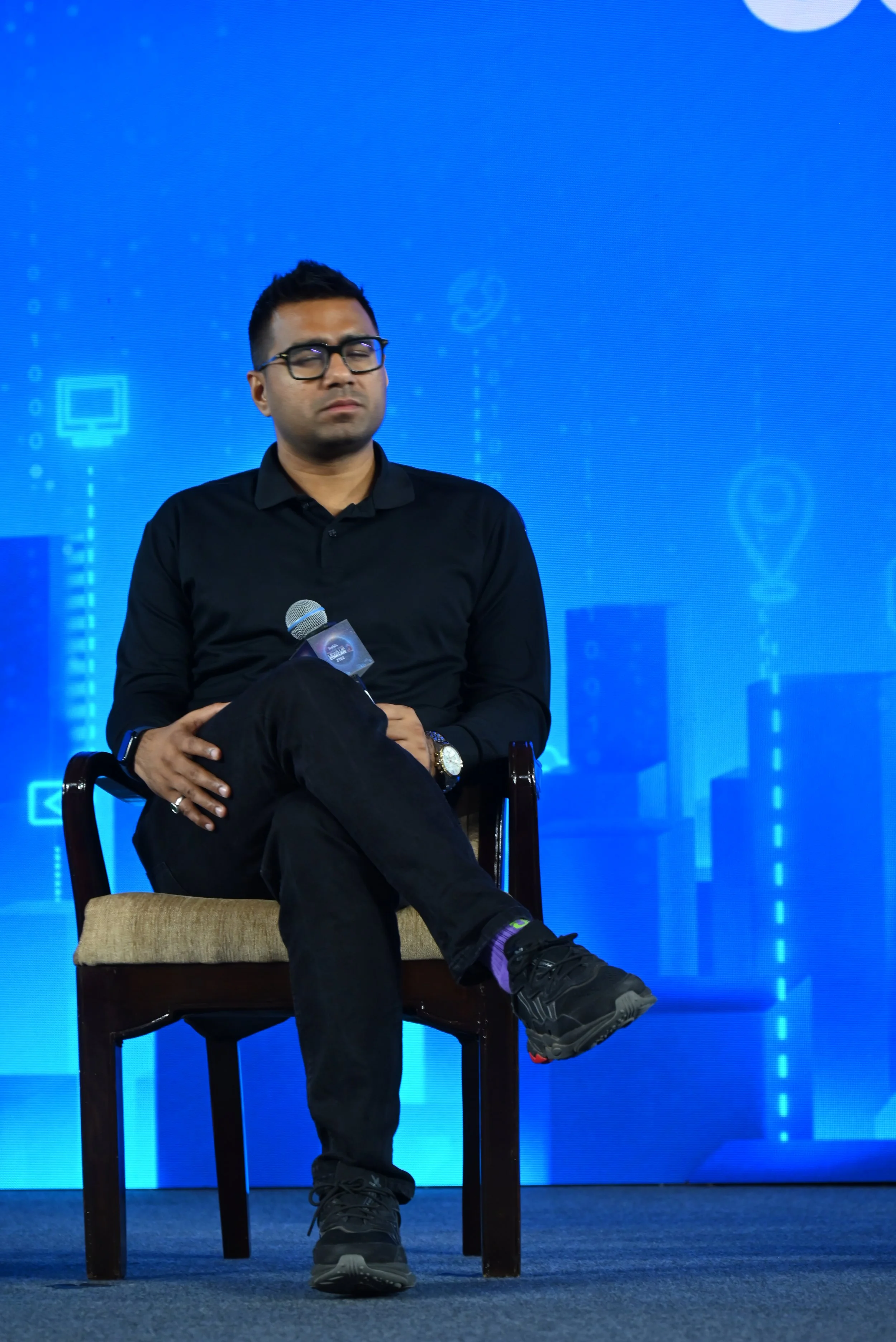 Man sitting on a chair with eyes closed, holding a microphone, wearing glasses, black shirt, black pants, and sneakers, on a stage with a blue digital-themed background.