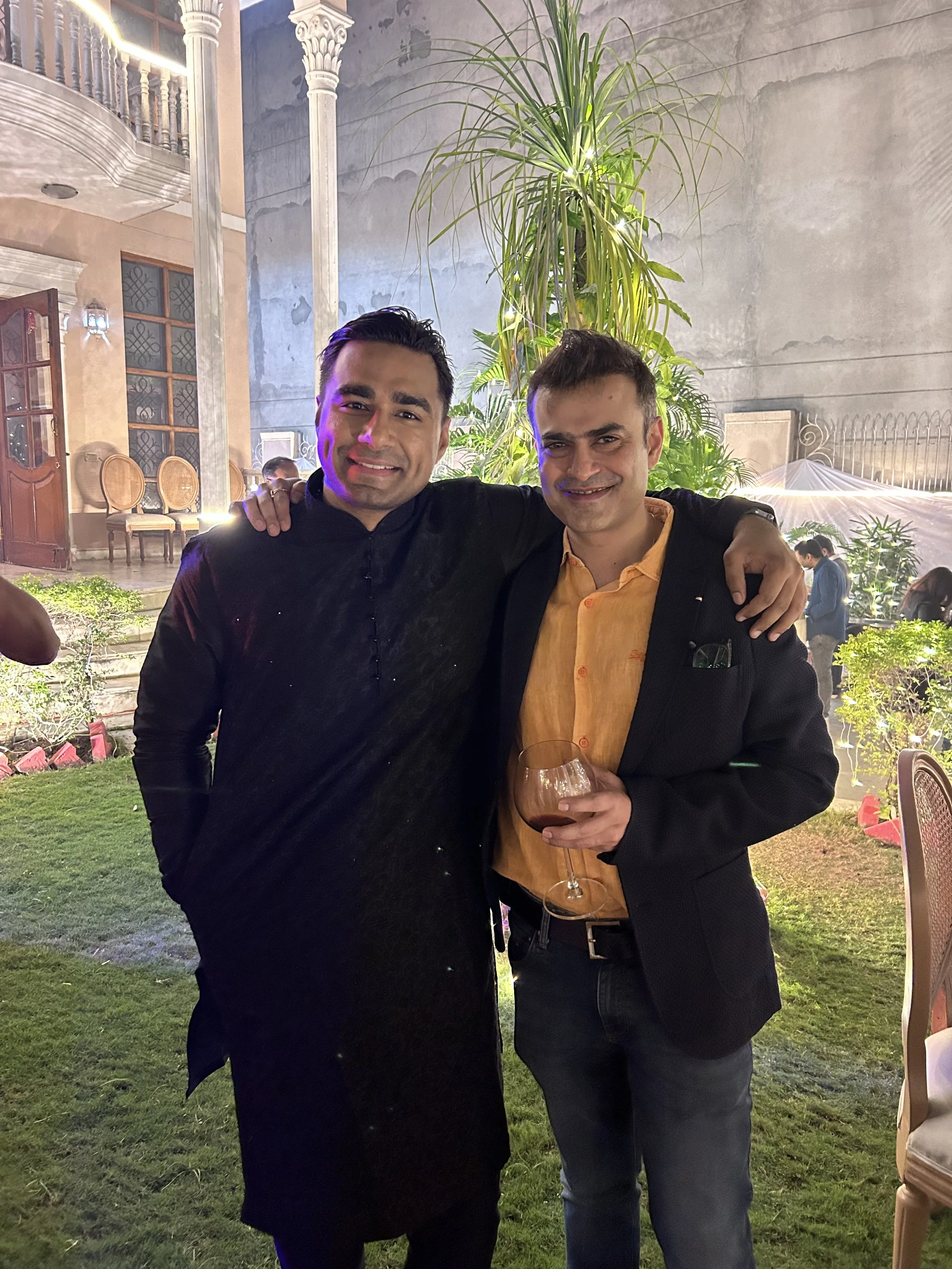 Two men standing outdoors at night, smiling, with one holding a glass of red wine. They have their arms around each other's shoulders. The background features greenery, a large plant, and an elegant building with arches and columns, illuminated with 