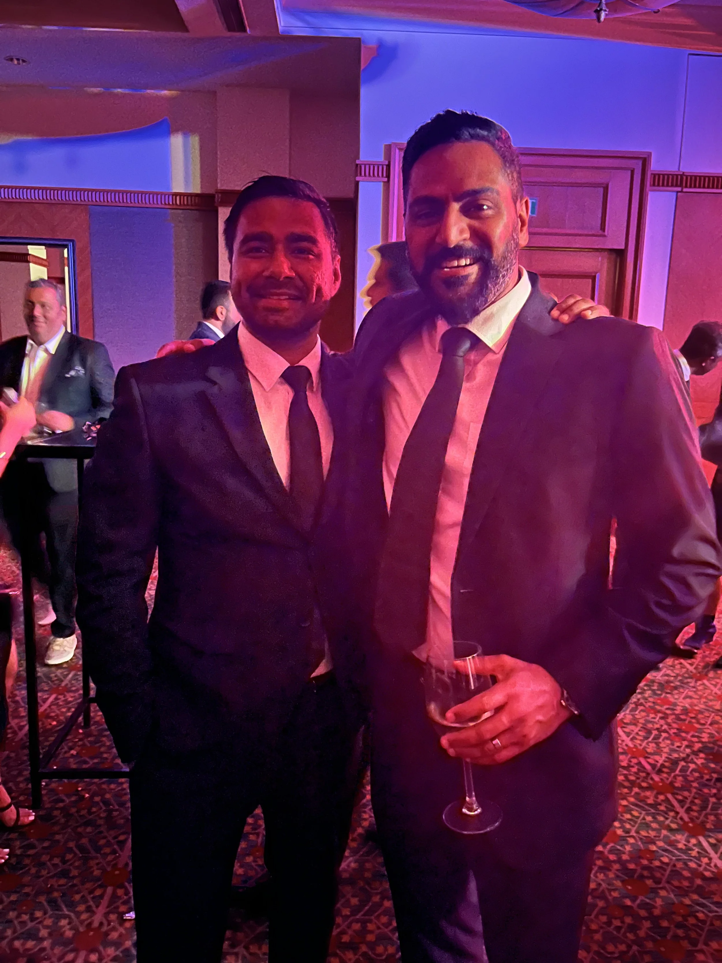 Two men in suits at a formal event, one holding a glass of wine, standing together and smiling.