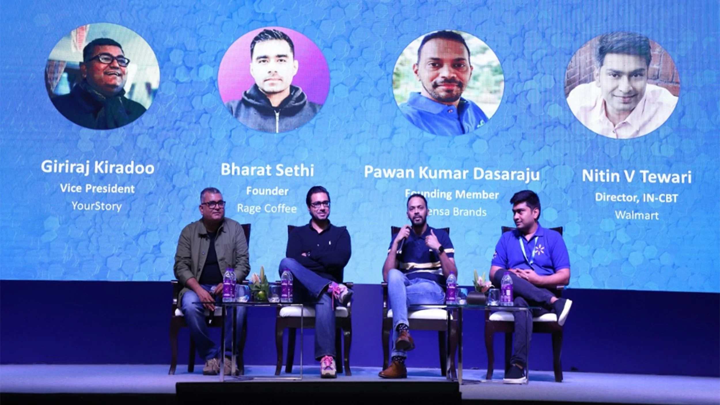 Four men sitting on chairs on stage during a panel discussion with a blue background displaying their names, titles, and photos.