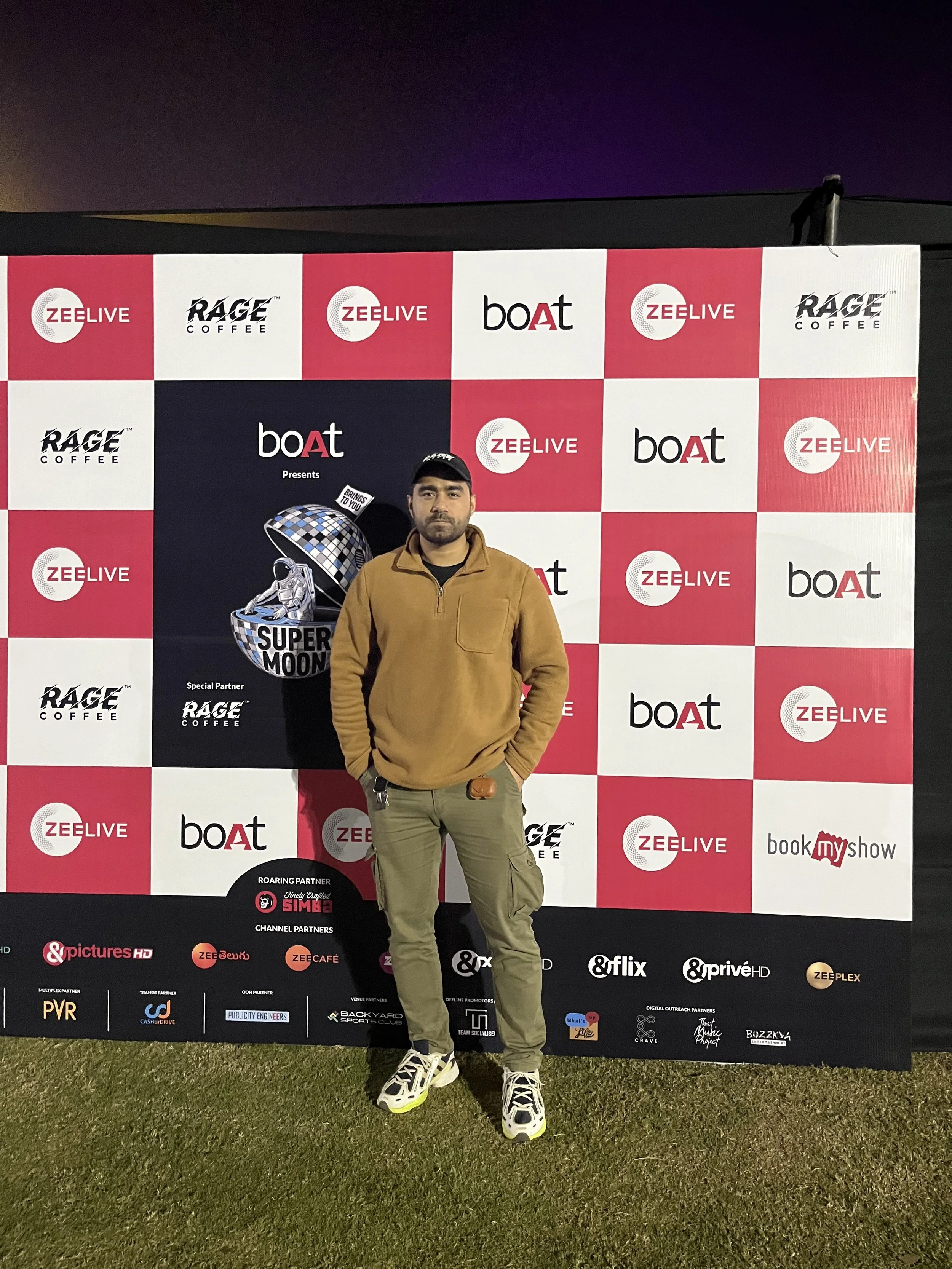 A man in a brown fleece jacket and green cargo pants standing in front of a promotional backdrop with various logos including Zee Live, boat, Rage Coffee, and others. The backdrop features an image of an astronaut and a moon.