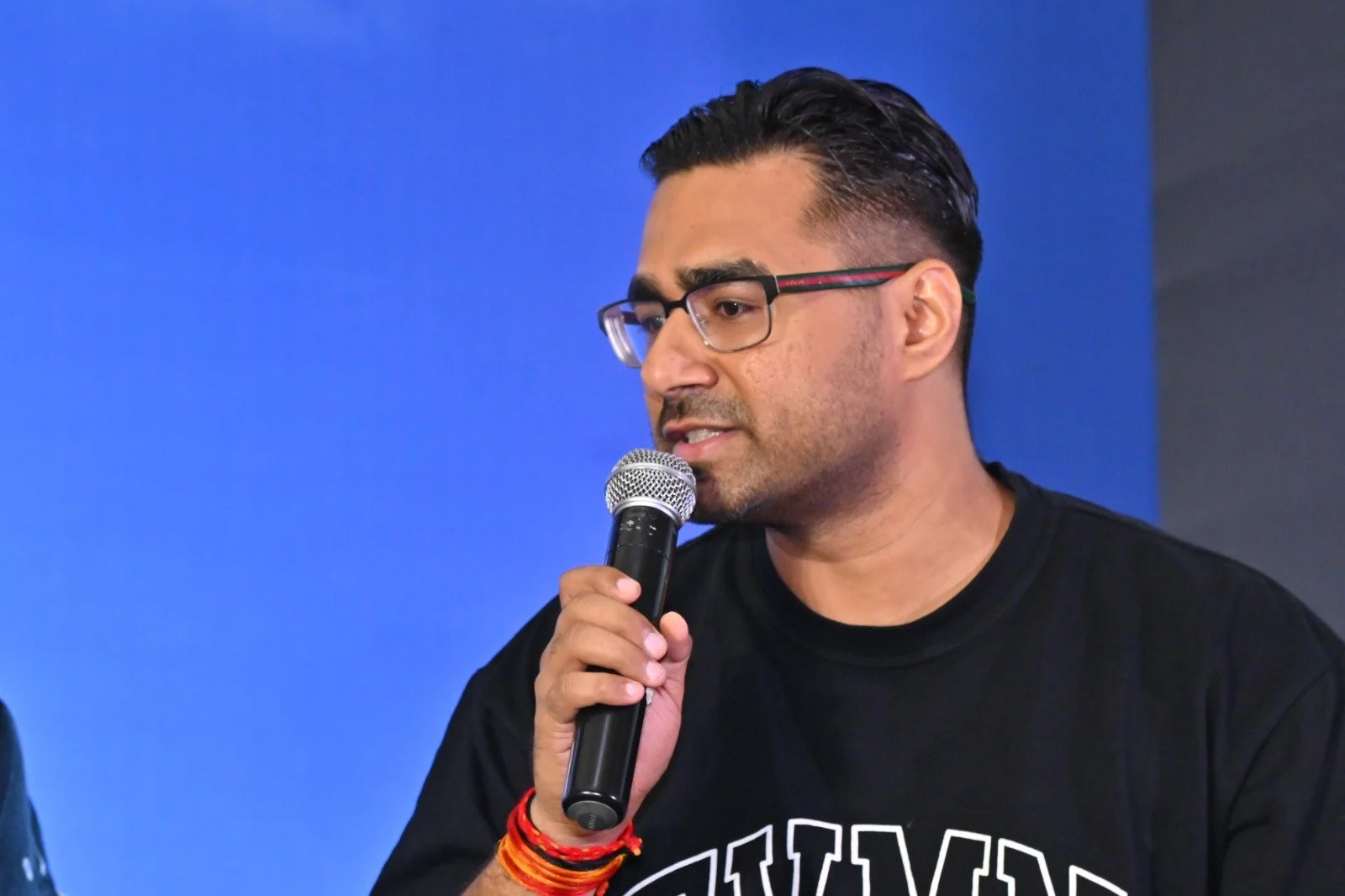 A man with dark hair, wearing glasses and a black T-shirt, speaks into a microphone against a blue background.