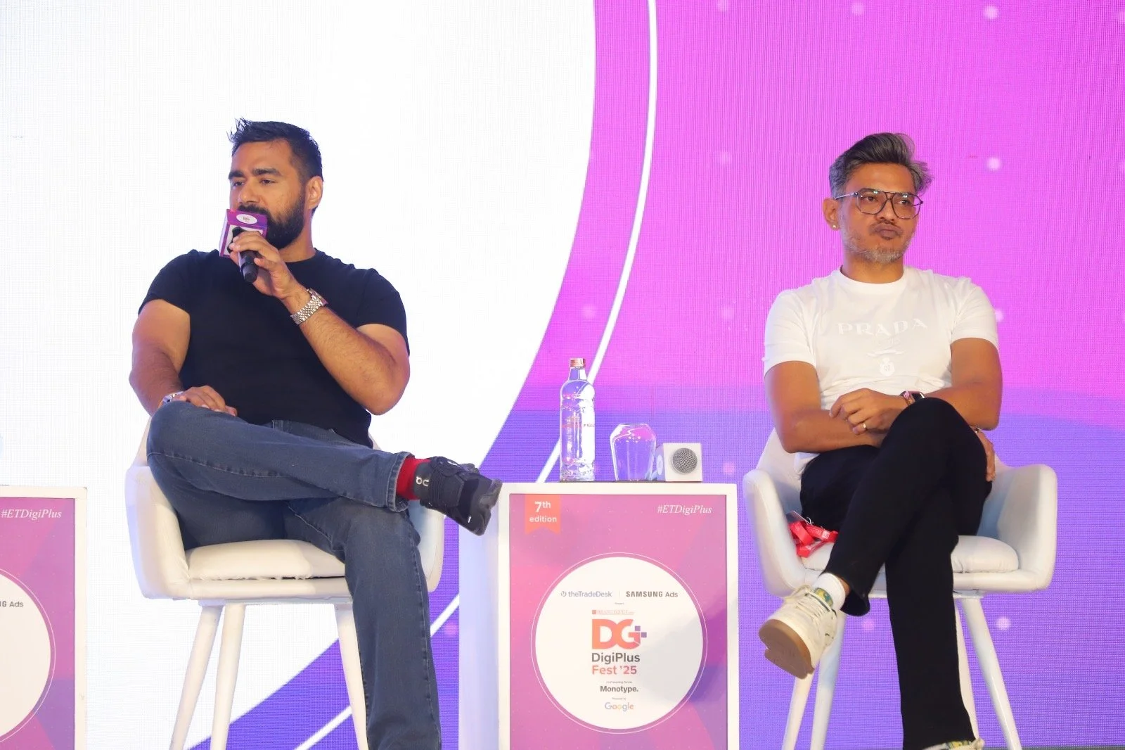 Two men seated on stage during the DigiPlus Fest '25, participating in a panel discussion. The man on the left is holding a microphone, wearing a black shirt and jeans, with his legs crossed. The man on the right is wearing a white Prada T-shirt, bla