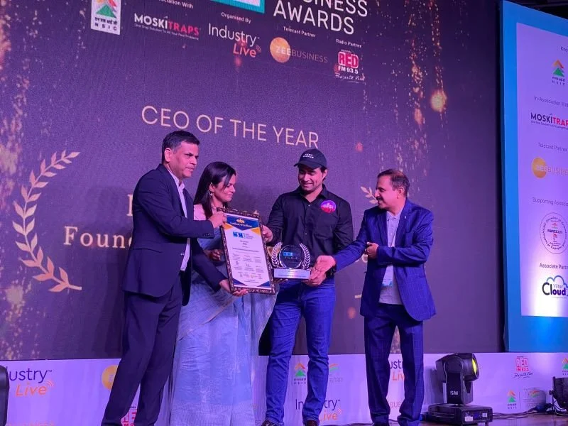 Four people on stage at an awards ceremony, with a purple backdrop displaying event logos and text that reads 'CEO of the Year.' One man is receiving an award and a framed certificate from another man, while a woman and a man stand beside them.