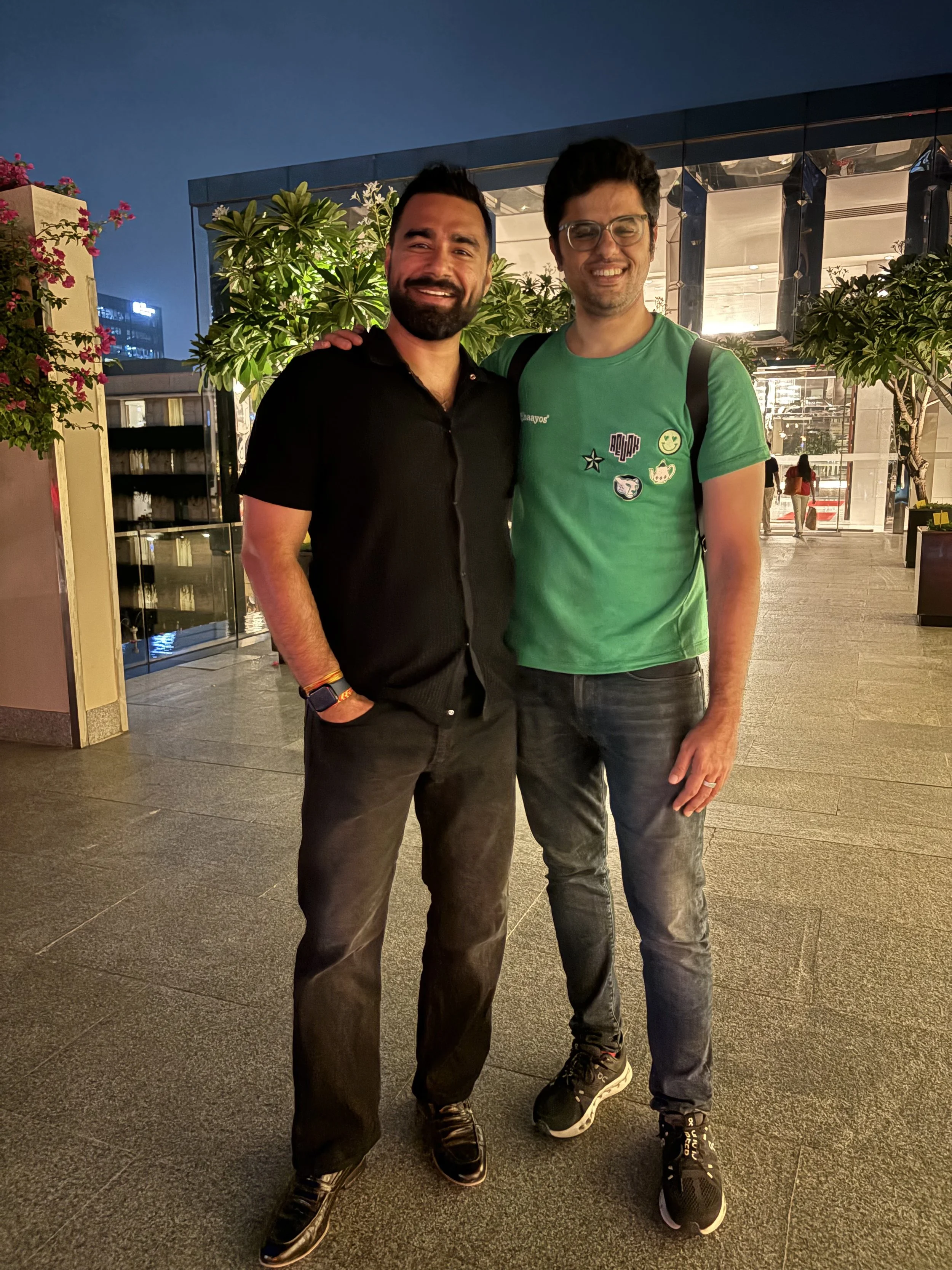 Two men standing together outdoors at night, smiling, with greenery and a modern building behind them.