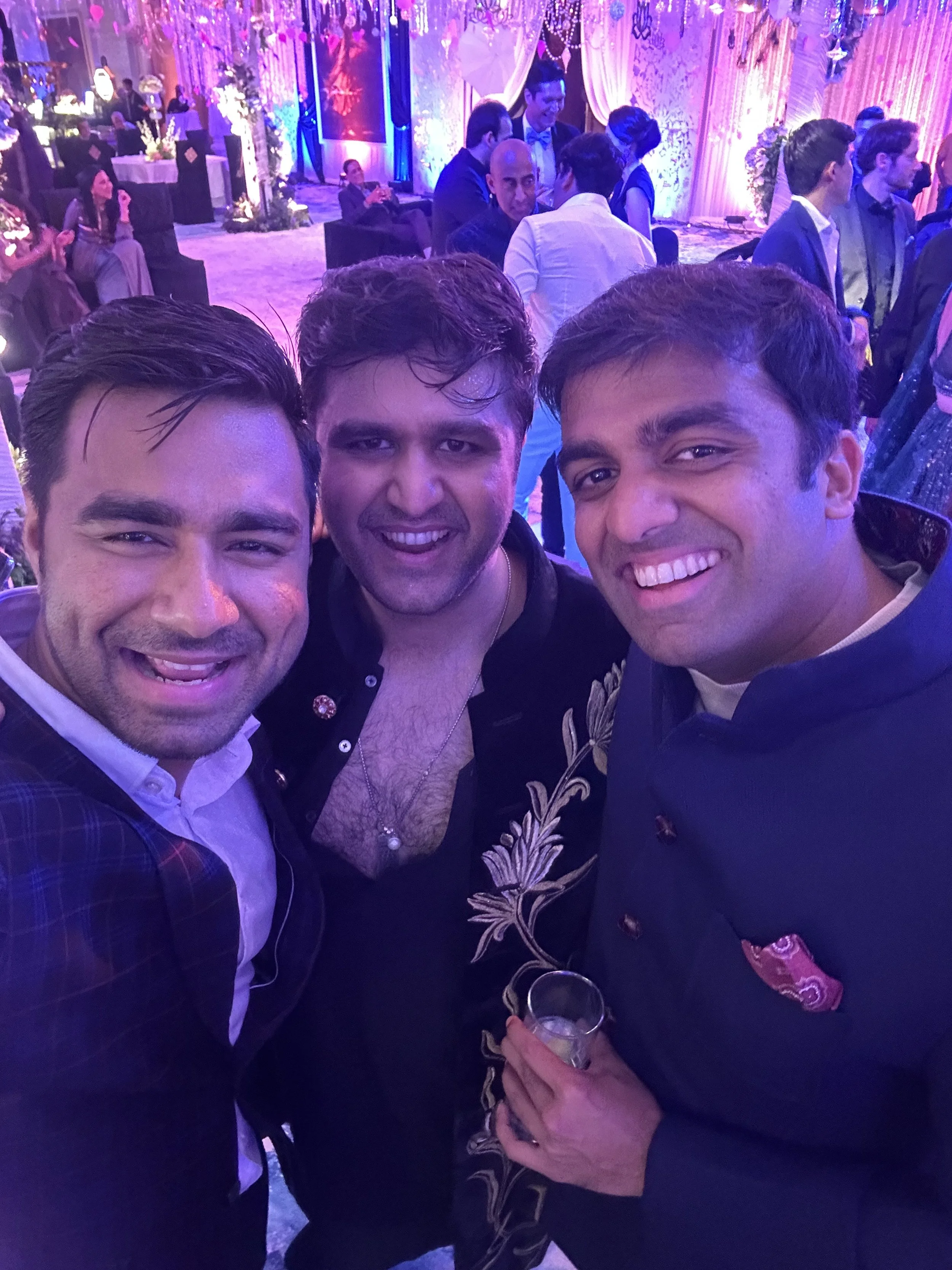Three men in formal attire smiling for a selfie at a festive indoor event with purple and pink lighting.