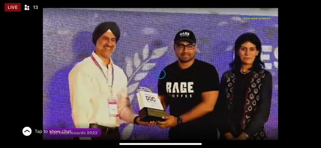 Three people on stage, one man in a white shirt and turban receiving an award from a man in a black 'RAGE Coffee' t-shirt, with a woman standing next to them, in front of a blue digital background.