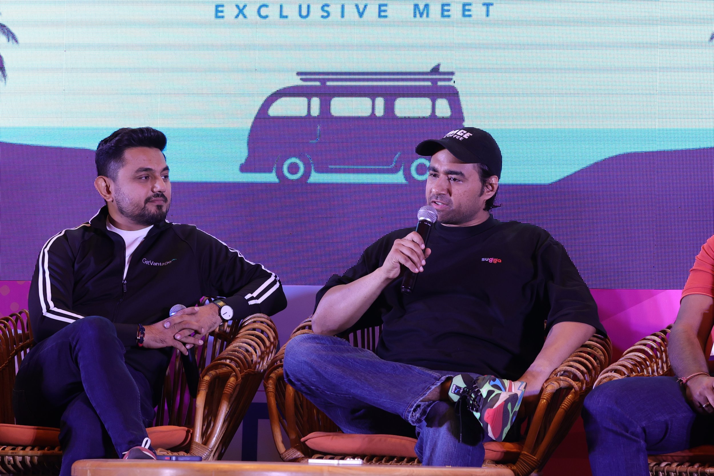Two men sitting on rattan chairs on stage during a panel discussion. The man on the left wears a black jacket with white stripes, and the man on the right wears a black t-shirt, a black cap, and colorful sneakers. The man on the right is speaking int