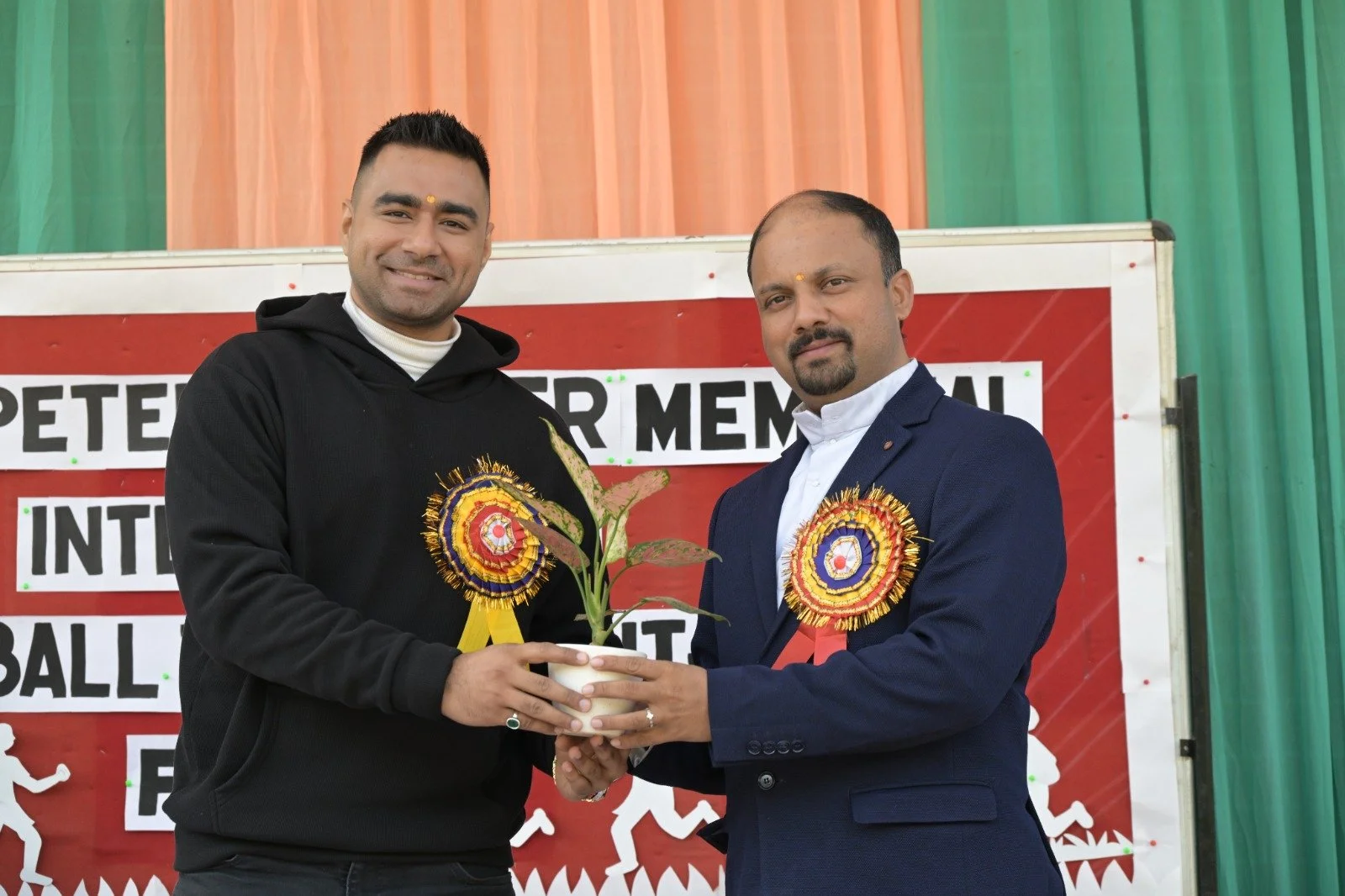 Two men standing in front of a decorated backdrop, exchanging a potted plant. Both men are dressed in formal attire with badge decorations on their chest, smiling at the camera.