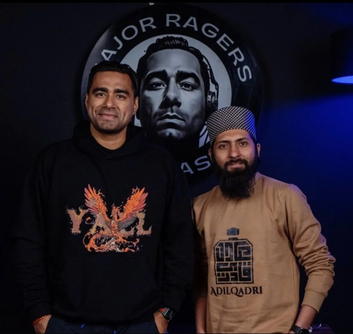 Two men standing in front of a wall with a logo featuring a man's face with headphones, with the text "Major Ragers" around the logo. One man is wearing a black hoodie with an orange embroidered phoenix and the word "YACL," and the other man is weari
