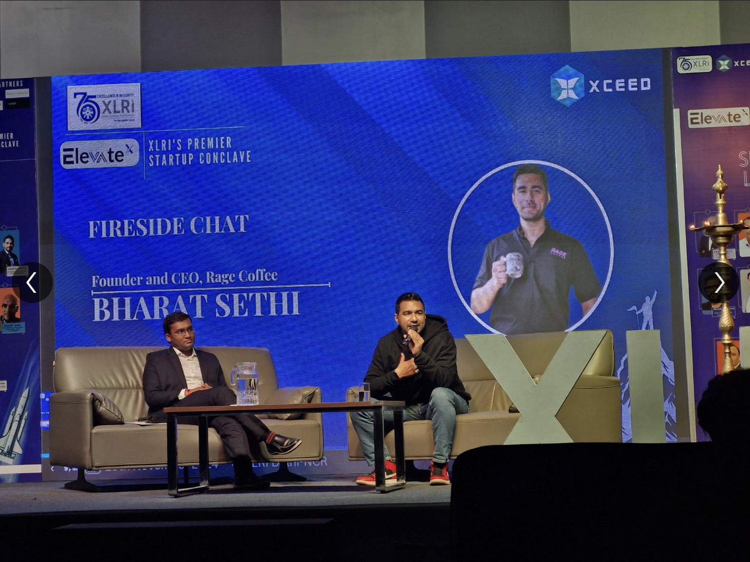 Two men sitting on sofas on a stage, participating in a fireside chat at the XLRI startup conclave. The background has a large blue screen with event branding, including the name Bharat Sethi and the company Rage Coffee.