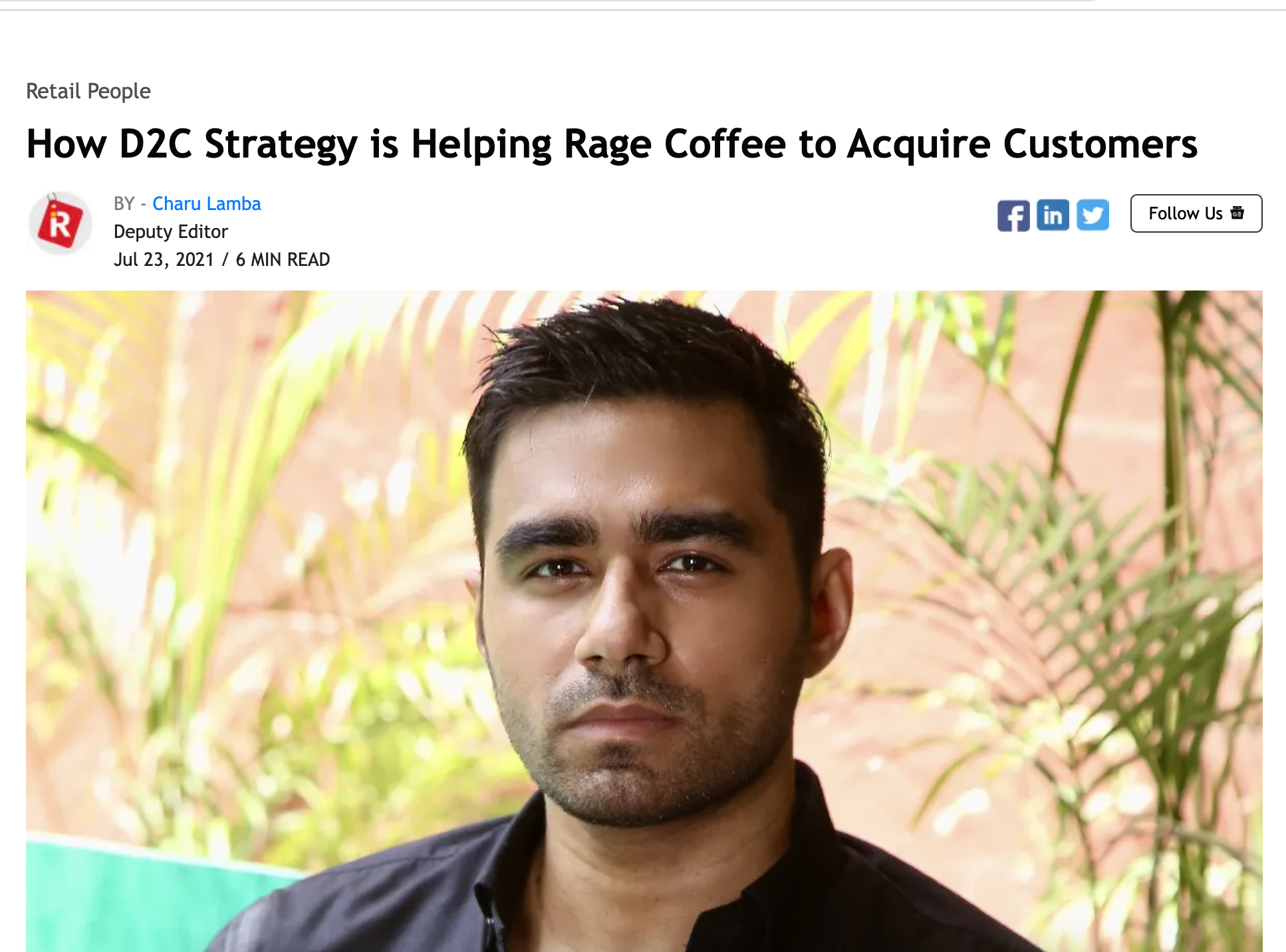 A man with short black hair and a serious expression, wearing a black shirt, standing outdoors with green foliage in the background.