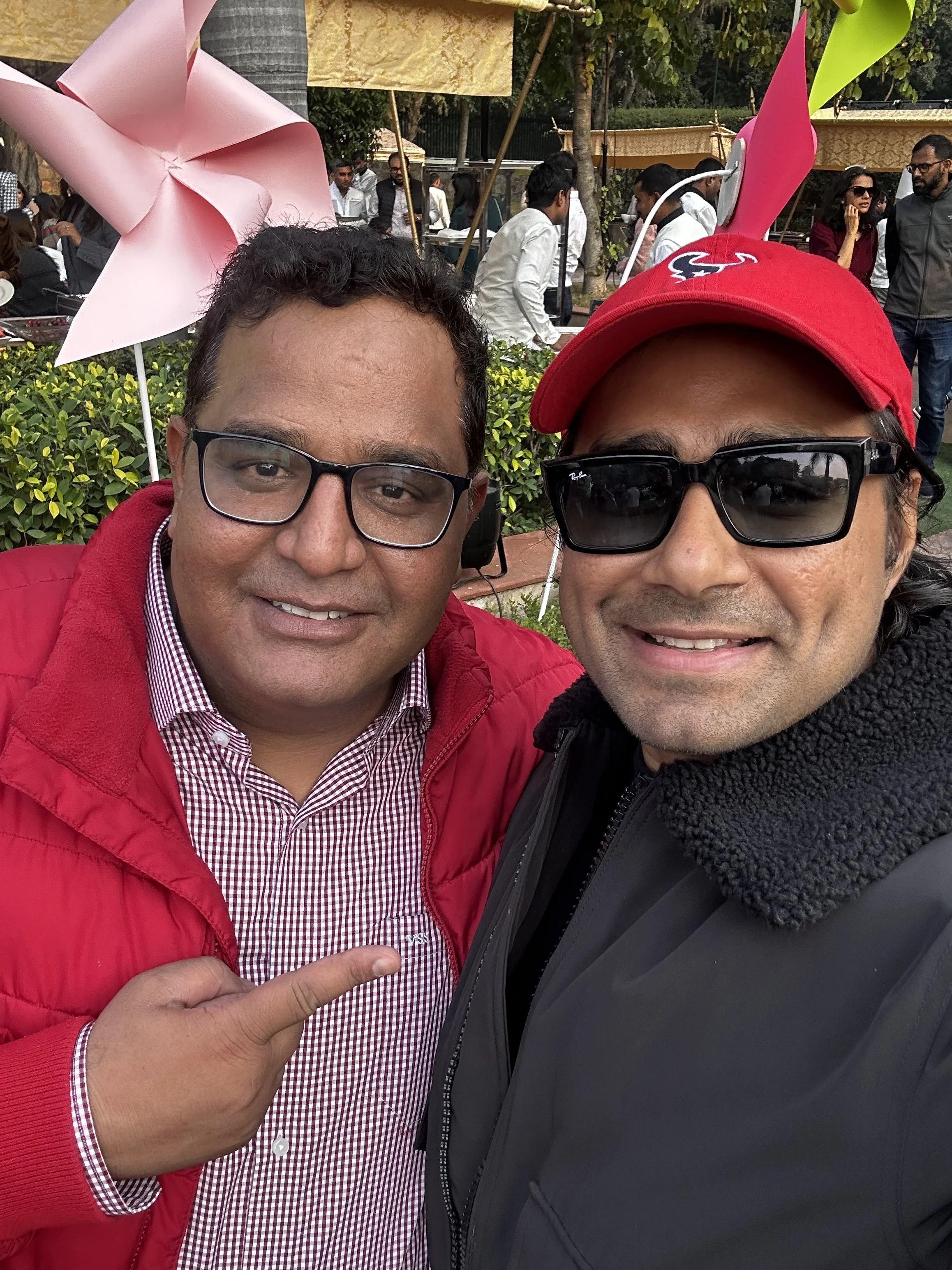 Two men taking a selfie outdoors at a festive event, one wearing a red jacket and the other wearing a black jacket with a red cap and sunglasses, with behind them pink pinwheel decorations and several other people in the background.
