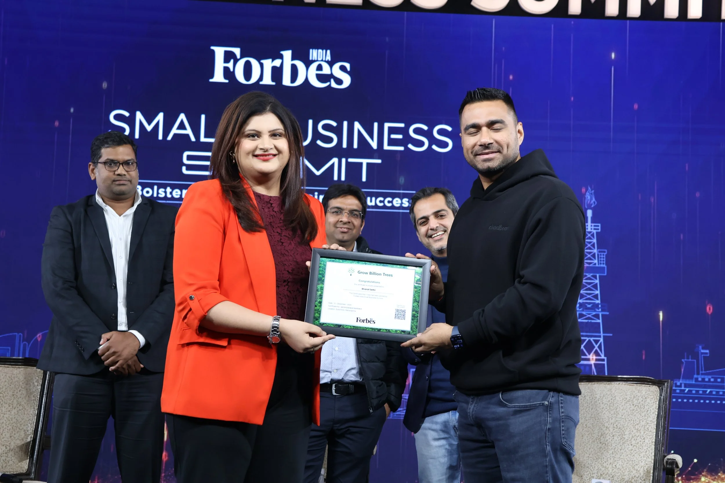 A group of five people, with two in the front holding a framed certificate, at a conference stage during the Forbes India Small Business Summit. The woman on the left with a red blazer is smiling, and the man on the right in a black hoodie is also sm
