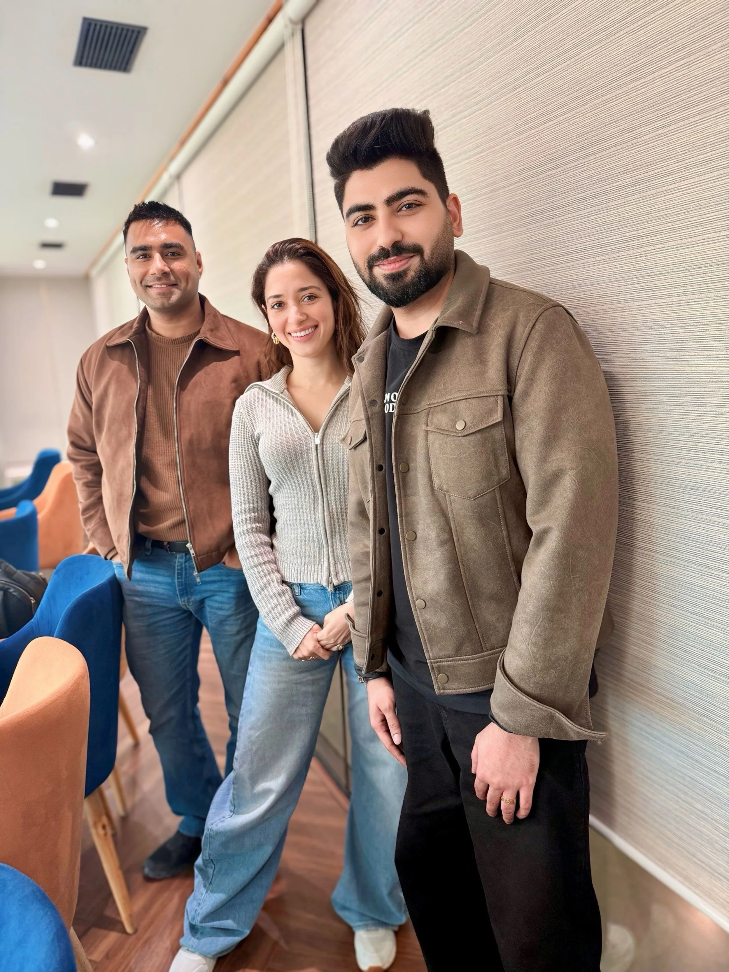 Three young adults standing indoors, smiling at the camera, with a wall and chairs in the background.