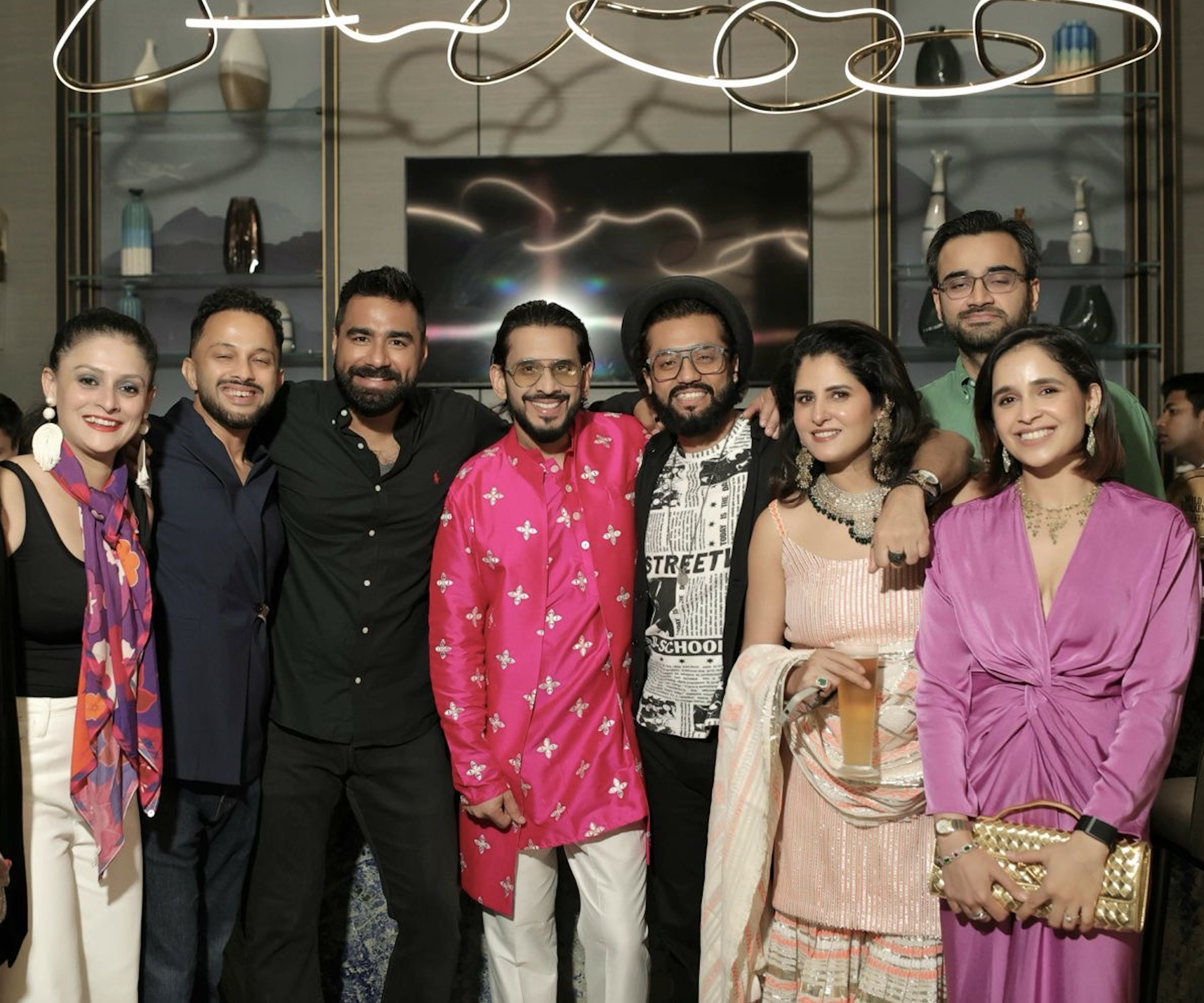 Group of eight people dressed in colorful party attire enjoying a social gathering in an upscale indoor setting, some holding drinks, smiling at the camera.