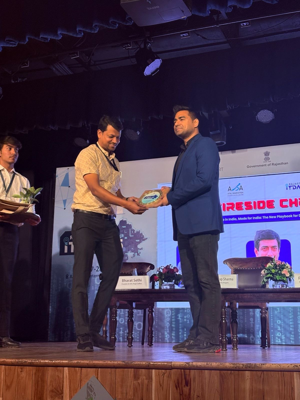A man in a blue blazer is receiving an award or plaque from another man in a beige shirt on a stage during a formal event, with a table and seats behind them and a screen displaying logos and event details in the background.