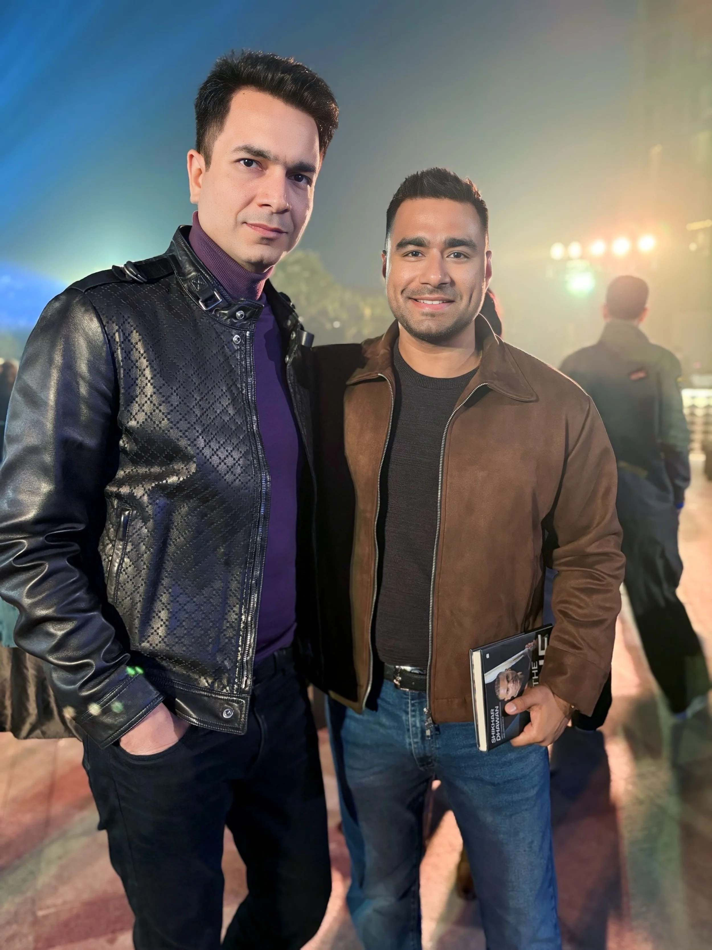 Two men standing outdoors at night, smiling at the camera. One wears a black leather jacket and the other a brown jacket, with a book or magazine in hand. Bright lights and blurred figures in background.