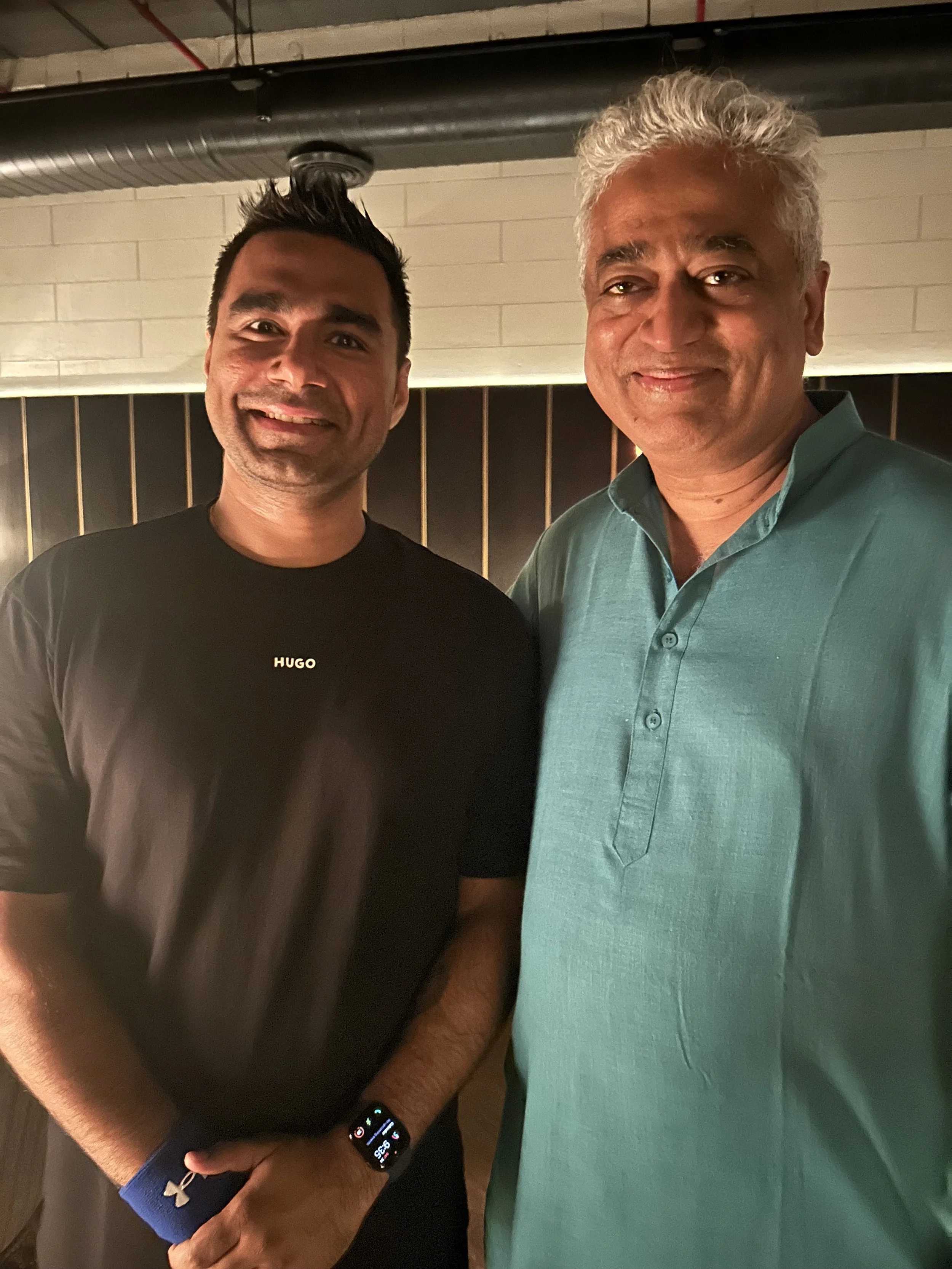 Two men standing together indoors, smiling at the camera. The man on the left wears a black t-shirt with "HUGO" on it and an Apple Watch, while the man on the right wears a teal collared shirt. The background features a beige brick wall and black pan