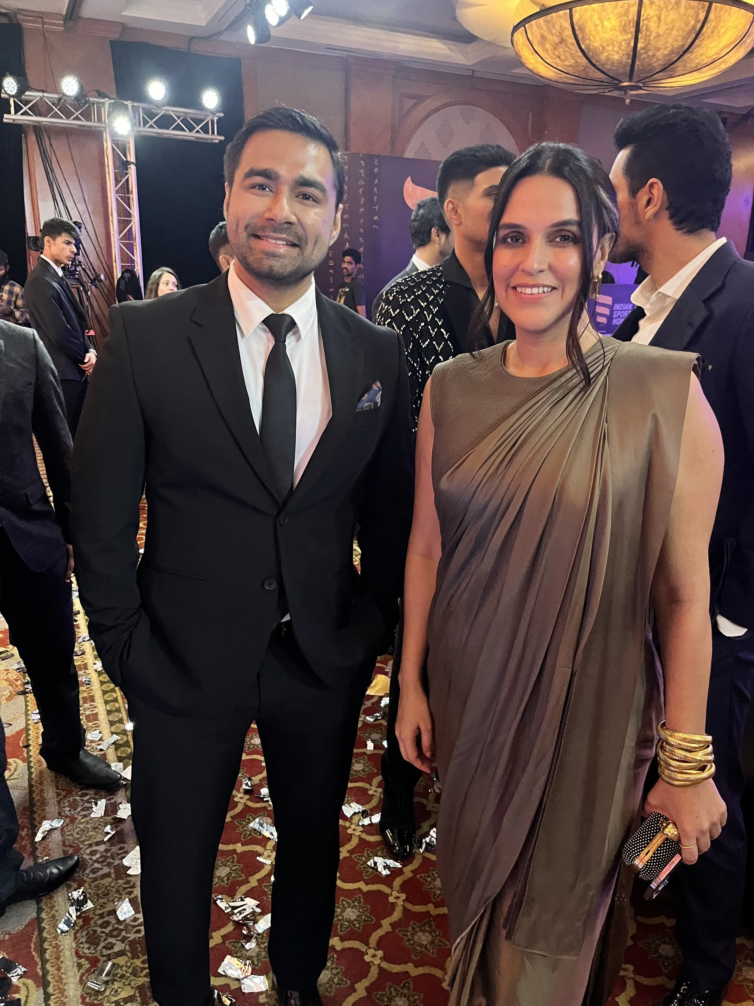 A man in a black suit and tie standing next to a woman in a taupe sari at a formal event, with other guests and a decorated banquet hall in the background.