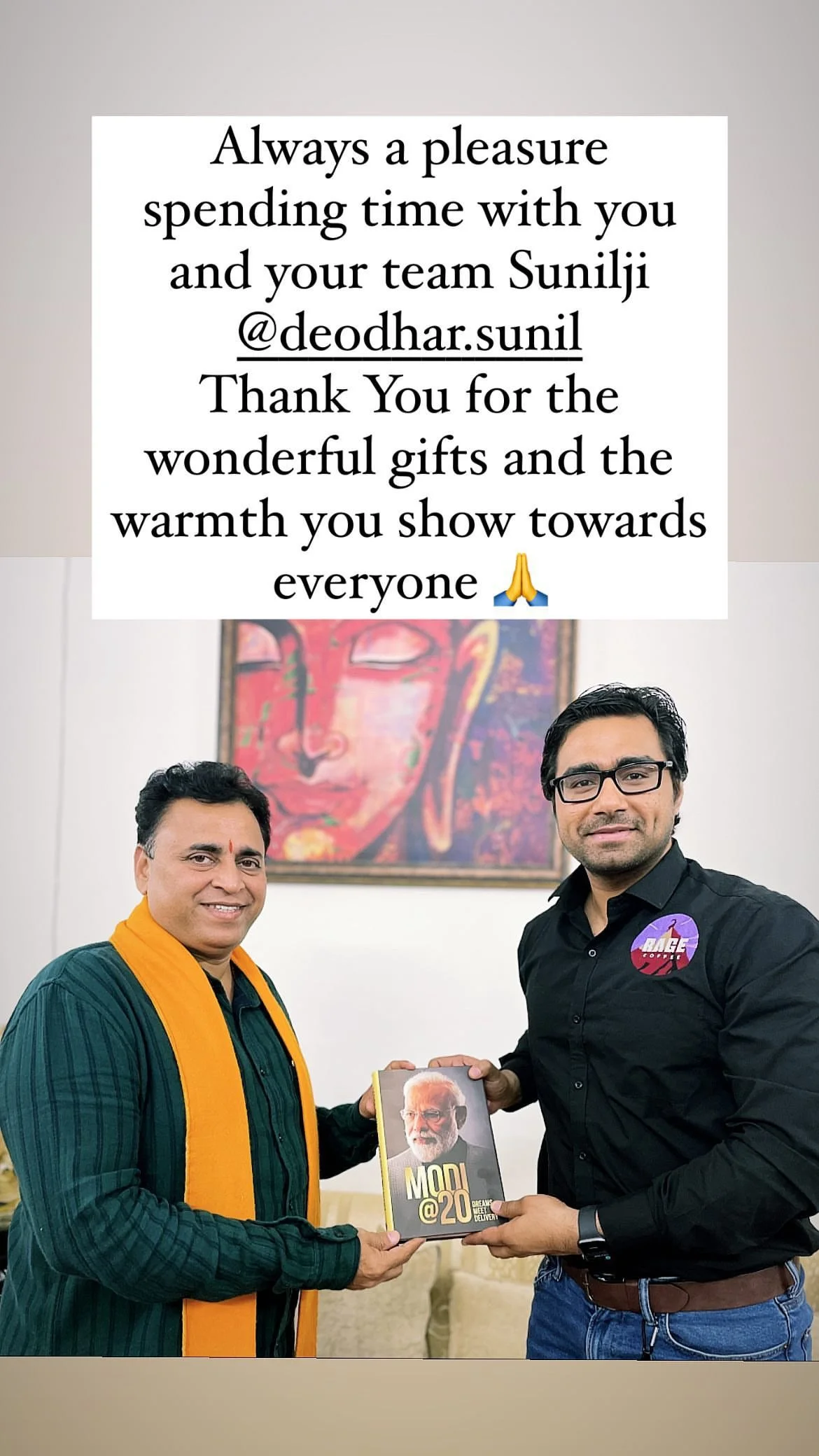 Two men are standing indoors, one is wearing a green kurta with an orange scarf and the other is dressed in a black shirt with a badge. They are holding a book titled 'MODI @ 20' together, smiling at the camera. A painting of Buddha's face is hanging