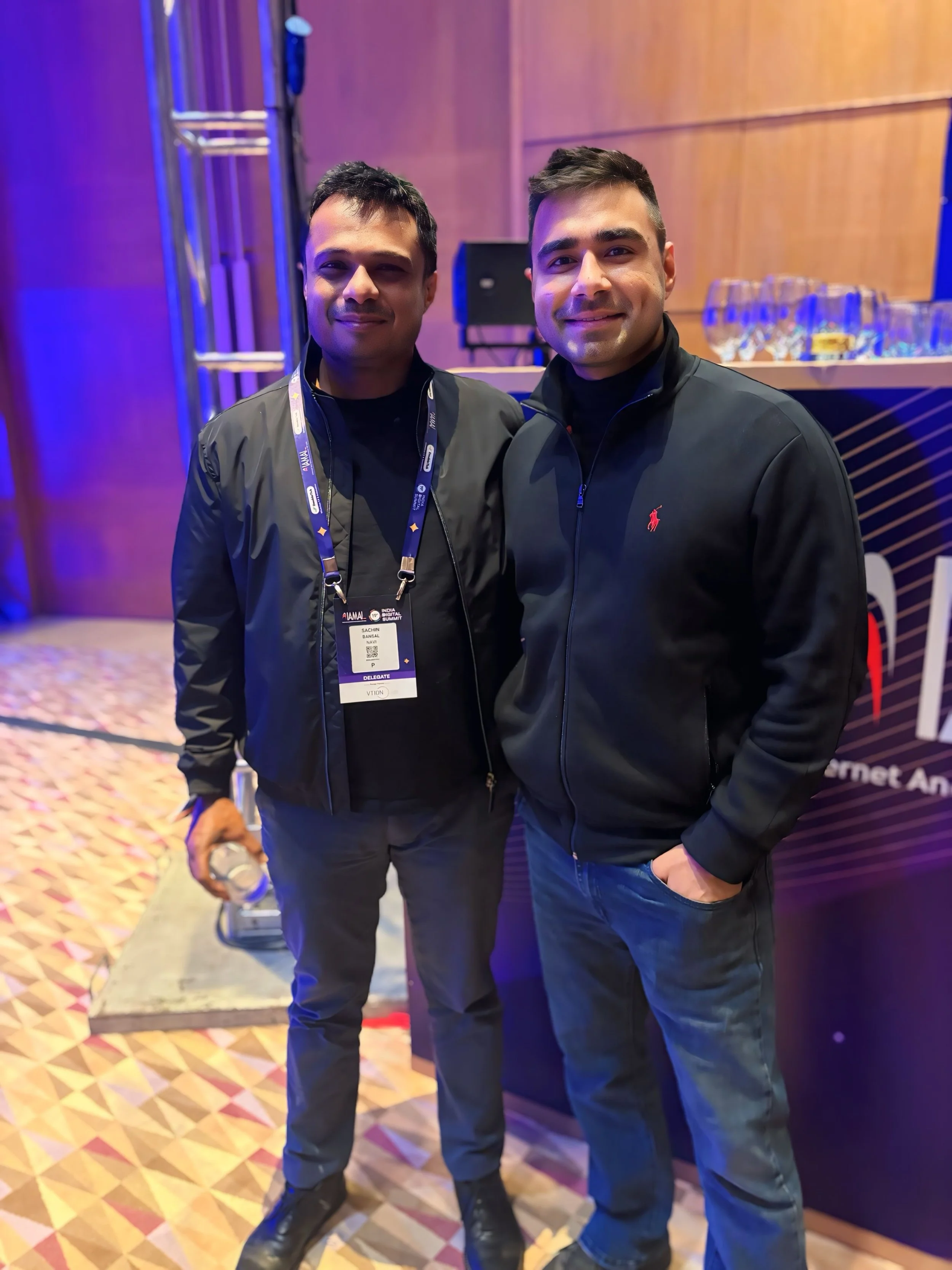 Two young men standing together at a conference or event, smiling for the photo. They are dressed in dark jackets, with one holding a water bottle. Behind them, there are glasses on a table and a colorful patterned carpet.