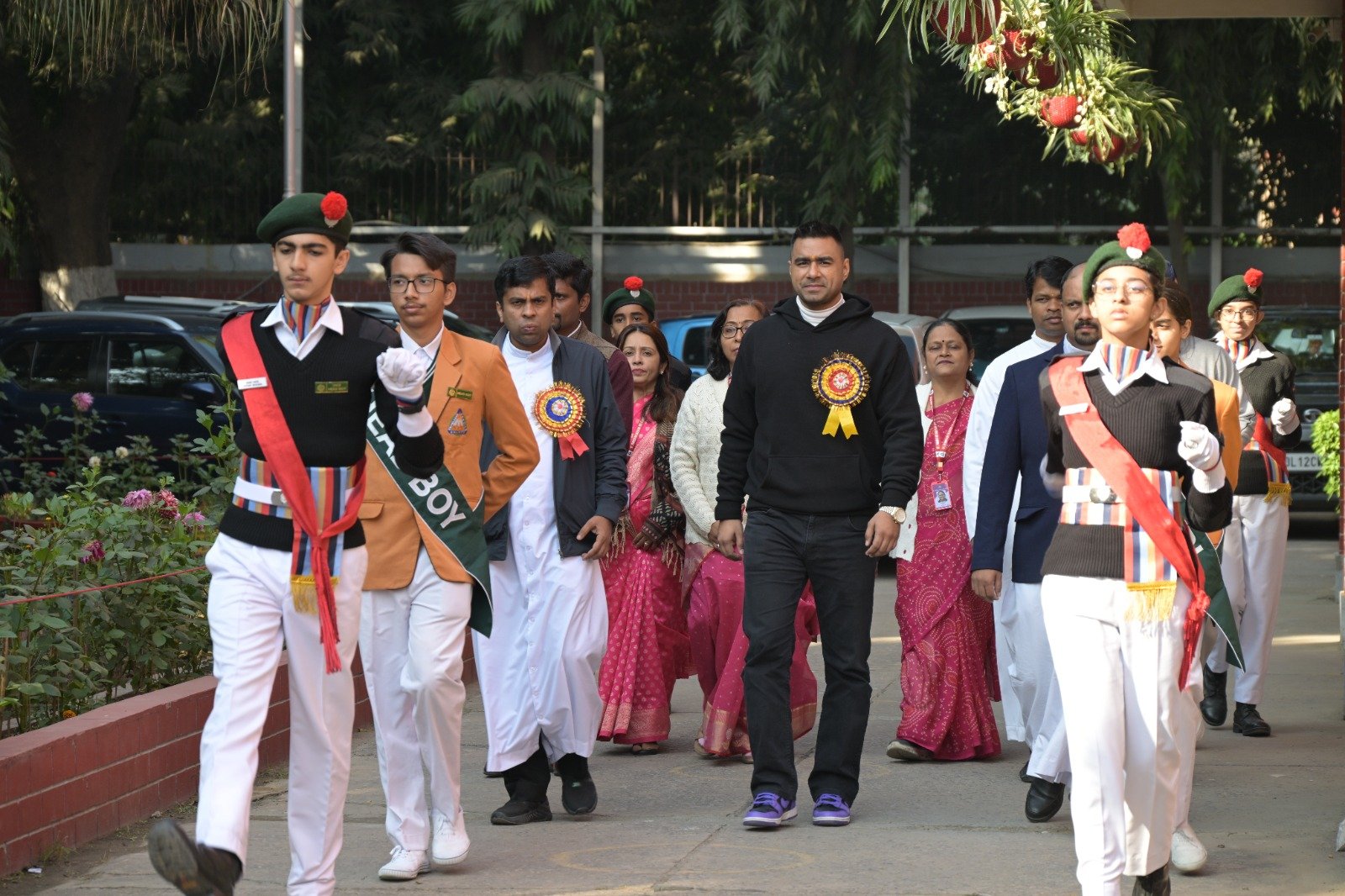 Group of people participating in a parade or procession, with some individuals dressed in formal or ceremonial attire, walking outdoors on a sidewalk with trees and parked cars in the background.