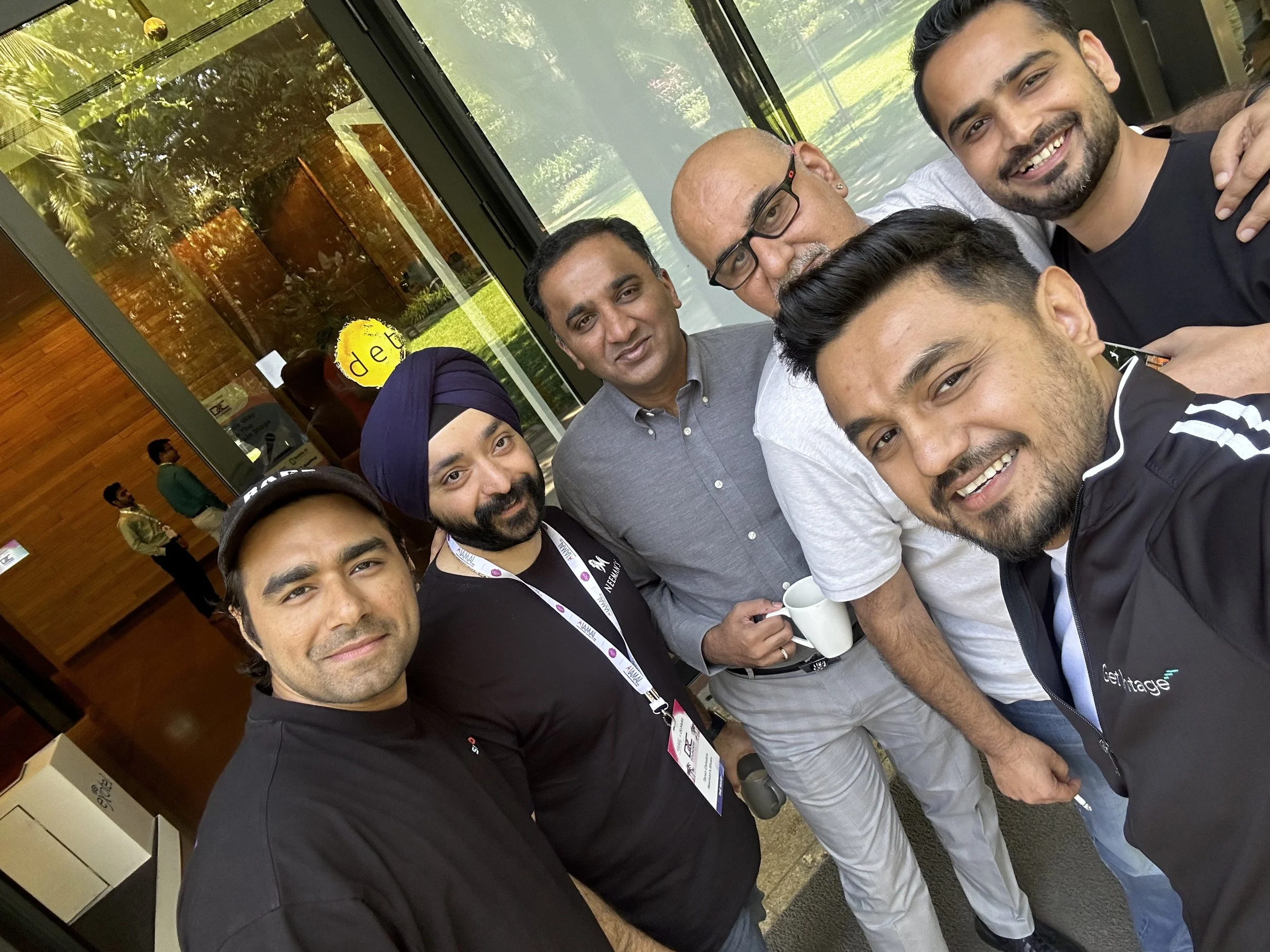 A group of seven men taking a selfie inside a building near large windows. The men are smiling and holding mugs. The background shows reflections and a glimpse of the outdoors.