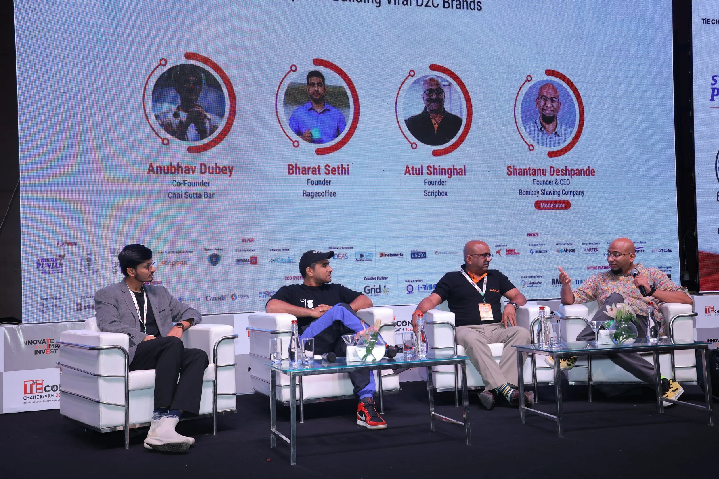 Panel discussion at a conference with four men sitting on stage in front of a large screen displaying profiles of four speakers. The profile pictures include names and titles: Anubhav Dubey, Co-Founder of Chai Sutta Bar; Bharat Sethi, Founder of Rage