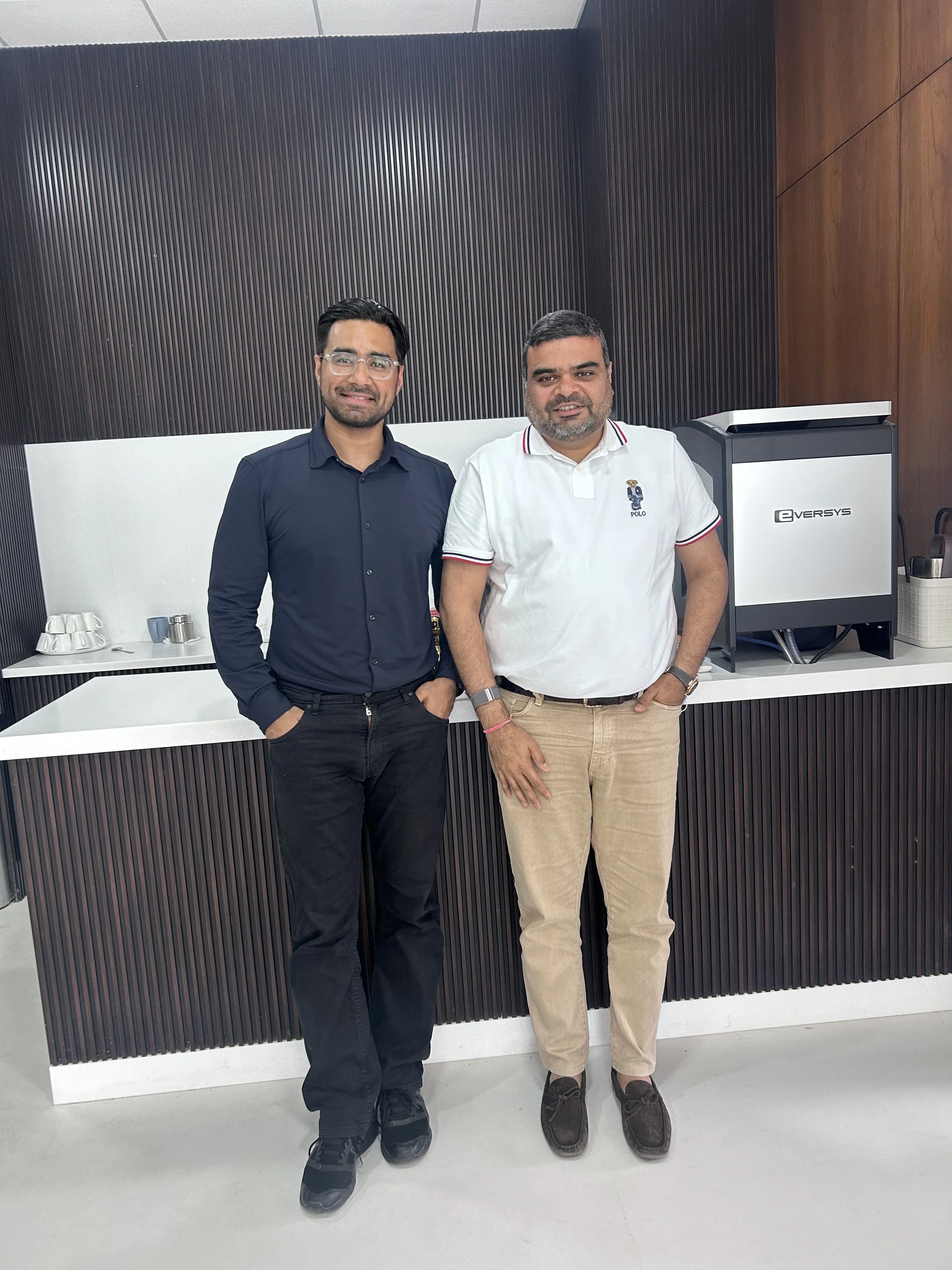 Two men standing side by side in an indoor setting, wearing casual and semi-formal attire, with a coffee machine and cups in the background.