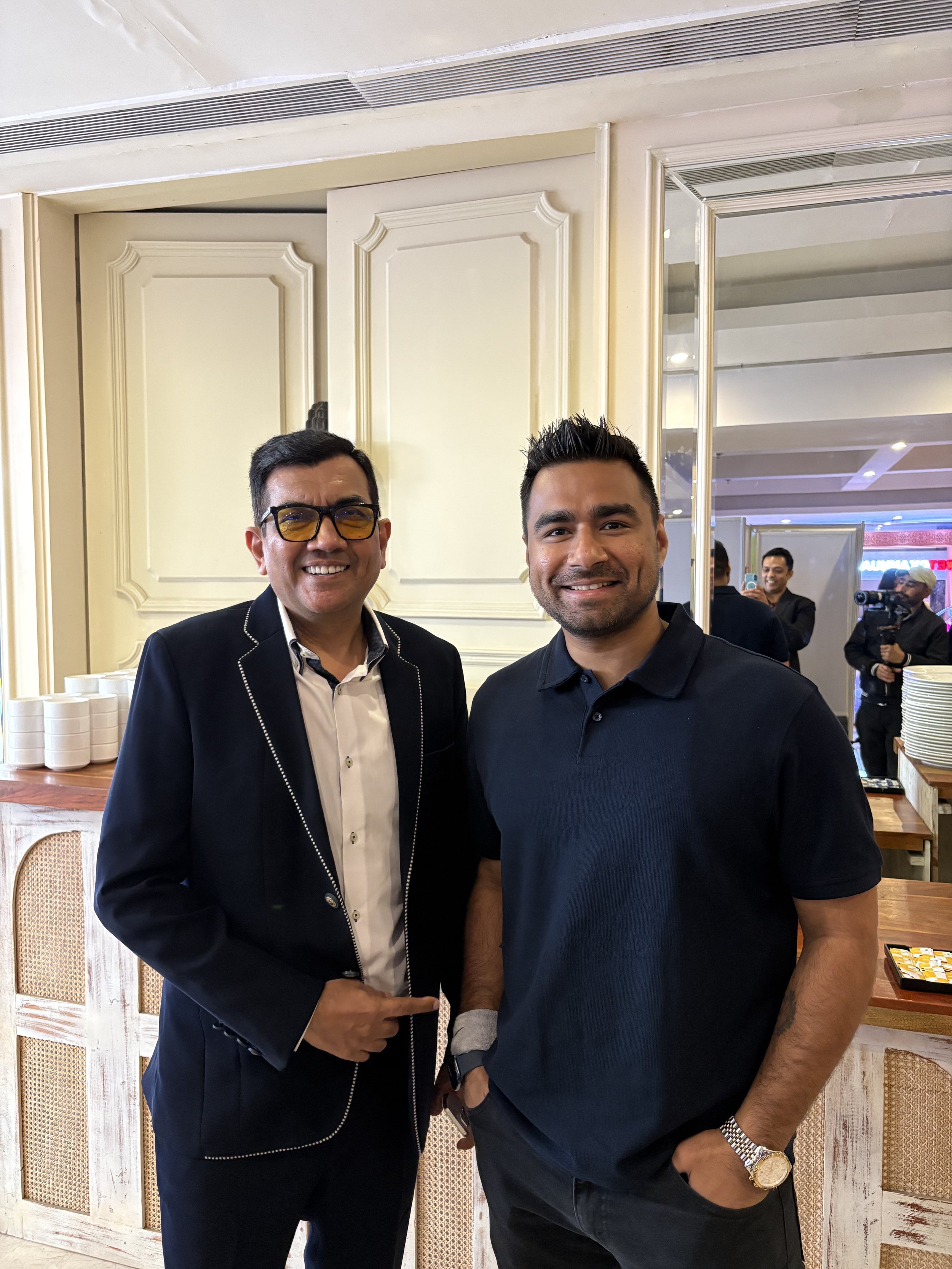 Two men standing next to each other smiling, in an indoor setting with a cream-colored interior and a mirror behind them. One man is wearing a black blazer with white piping, white shirt, and yellow-tinted glasses. The other man is dressed in a black