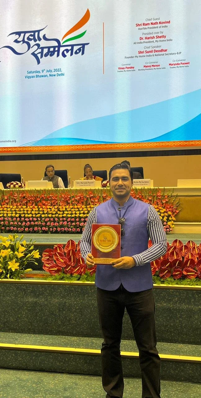 A man standing on a stage holding an award plaque, with a colorful flower arrangement surrounding him. In the background, there are three people sitting at a panel table with nameplates, behind a large screen displaying a logo and text about an event