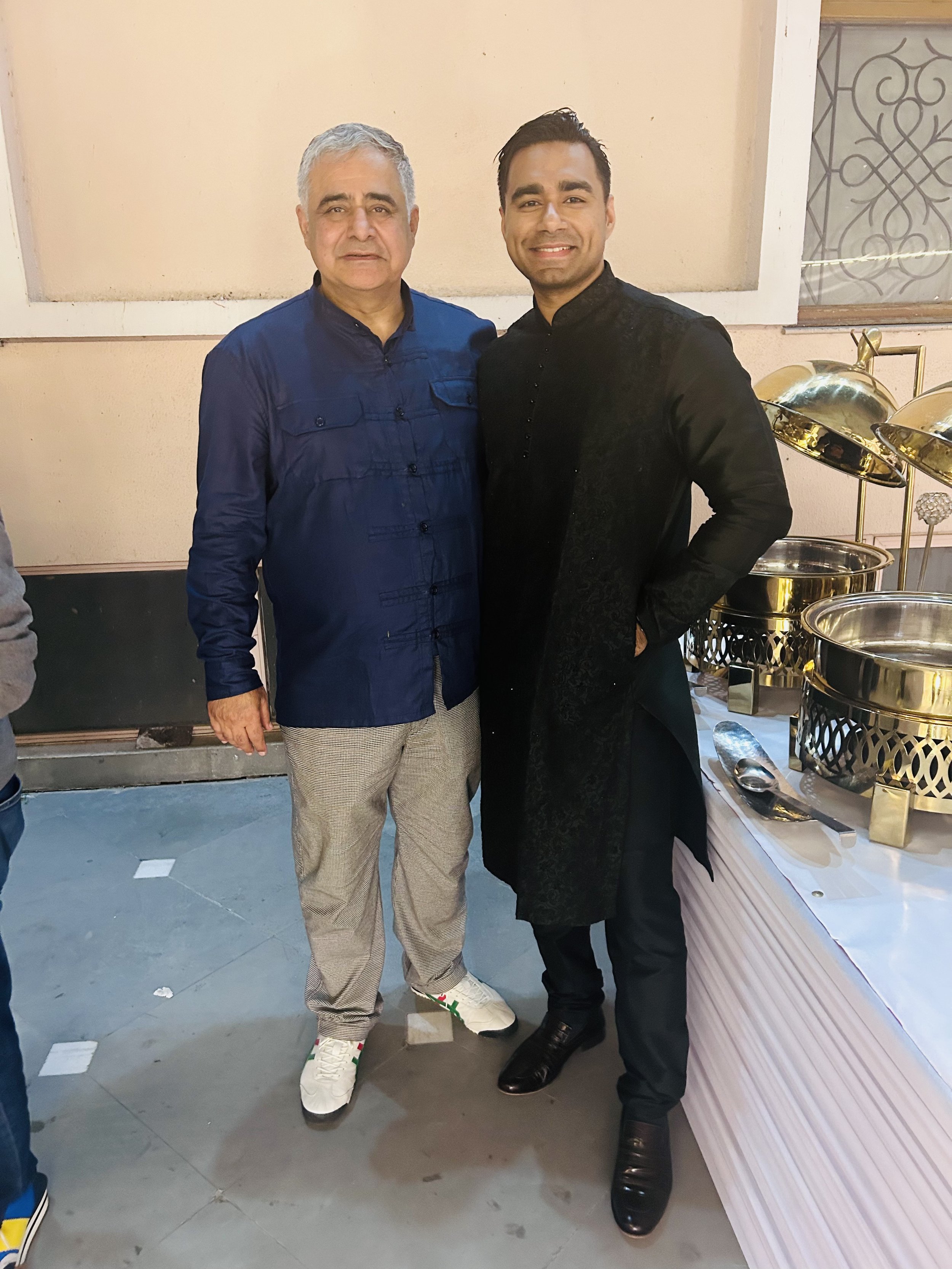 Two men standing side by side, smiling at the camera, at a buffet table with chafing dishes. One man is wearing a blue shirt and light-colored trousers, the other in traditional black attire. The setting appears to be a celebration or party.
