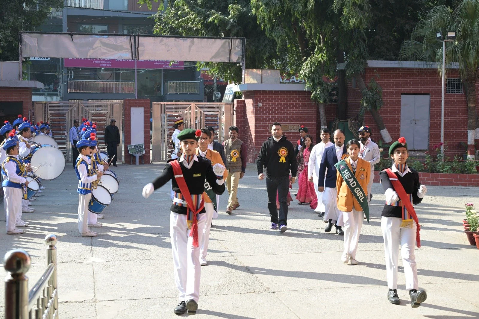 Students and teachers participating in a parade at school, with students in uniform and holding a banner that reads 'HEAD GIRL,' and a marching band of students in uniform playing drums.