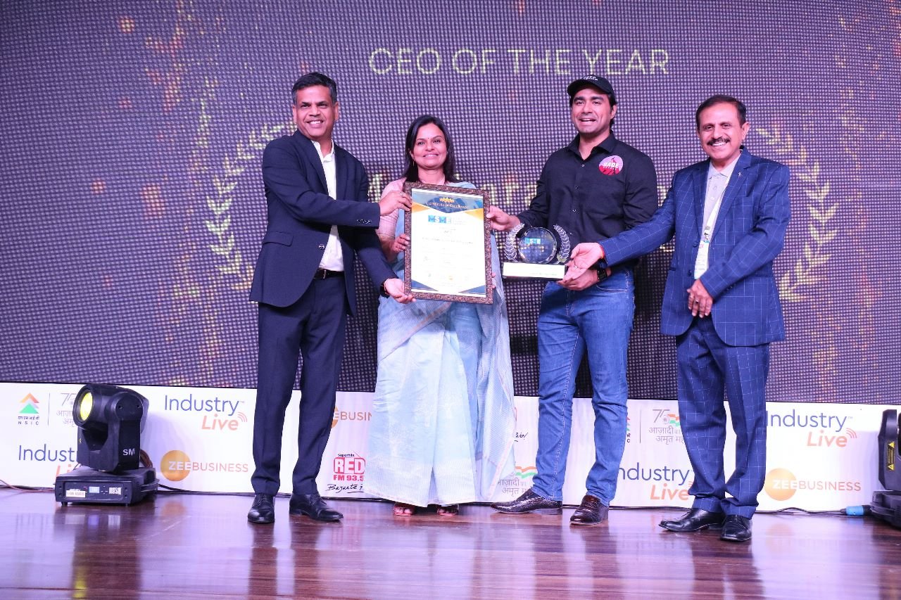 A group of four people on stage during an award ceremony. One person is holding a framed certificate, man next to her is holding a trophy and a transparent globe. The background displays the text "CEO OF THE YEAR" and is decorated with laurel designs