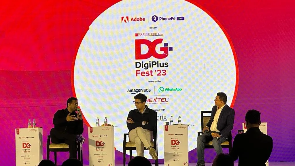 A panel discussion at DigPlus Fest 2023 with four men on stage, three seated and one speaking into a microphone. The backdrop displays event branding, including the DigPlus Fest 23 logo and sponsor logos like Adobe, Amazon Ads, WhatsApp, and others.