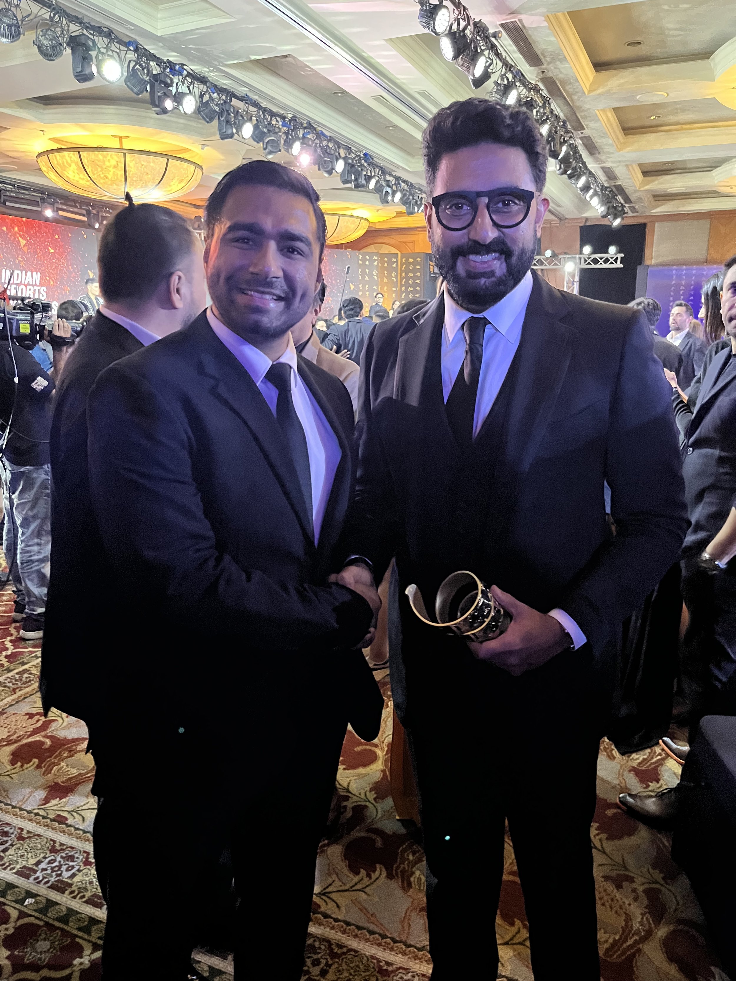 Two men in black suits shaking hands at an award ceremony, one holding a trophy, with a decorated banquet hall and stage in the background.