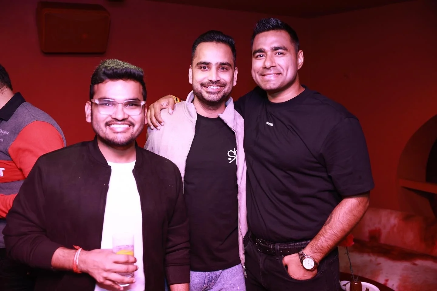 Three men smiling and standing together at a social gathering, in front of a red wall. The man on the left is wearing glasses and holding a drink, the middle man has his arm around the others, and the man on the right is wearing a black T-shirt and a