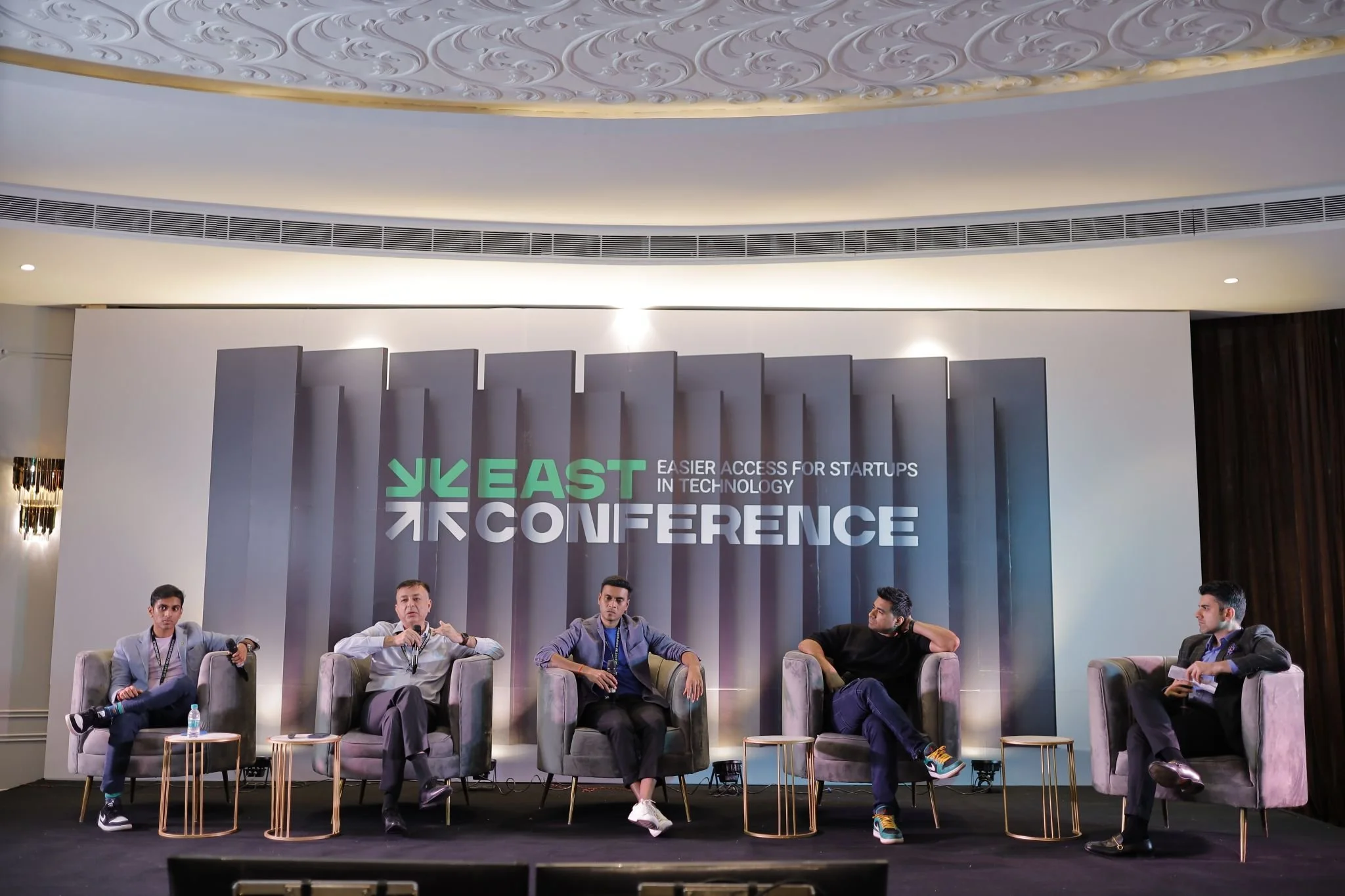 Five men seated on stage in armchairs during a conference with a backdrop that says 'EAST CONFERENCE' and 'EASIER ACCESS FOR STARTUPS IN TECHNOLOGY'.