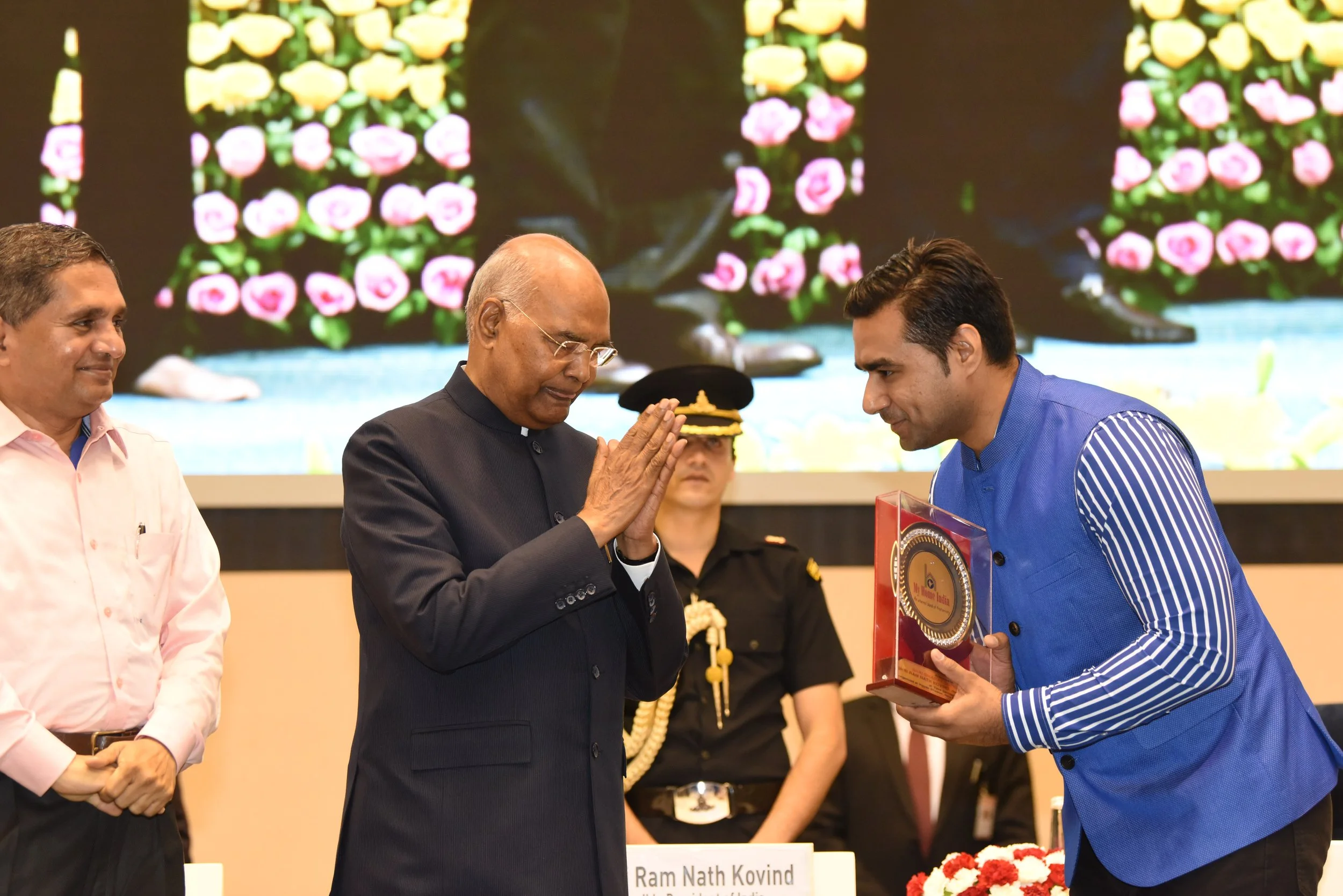 A man in a blue traditional Indian kurta receiving an award in a clear plastic case from a senior man in a black suit who is showing respect with folded hands during an indoor event. Two other men, one in a pink shirt and khaki pants and another in u