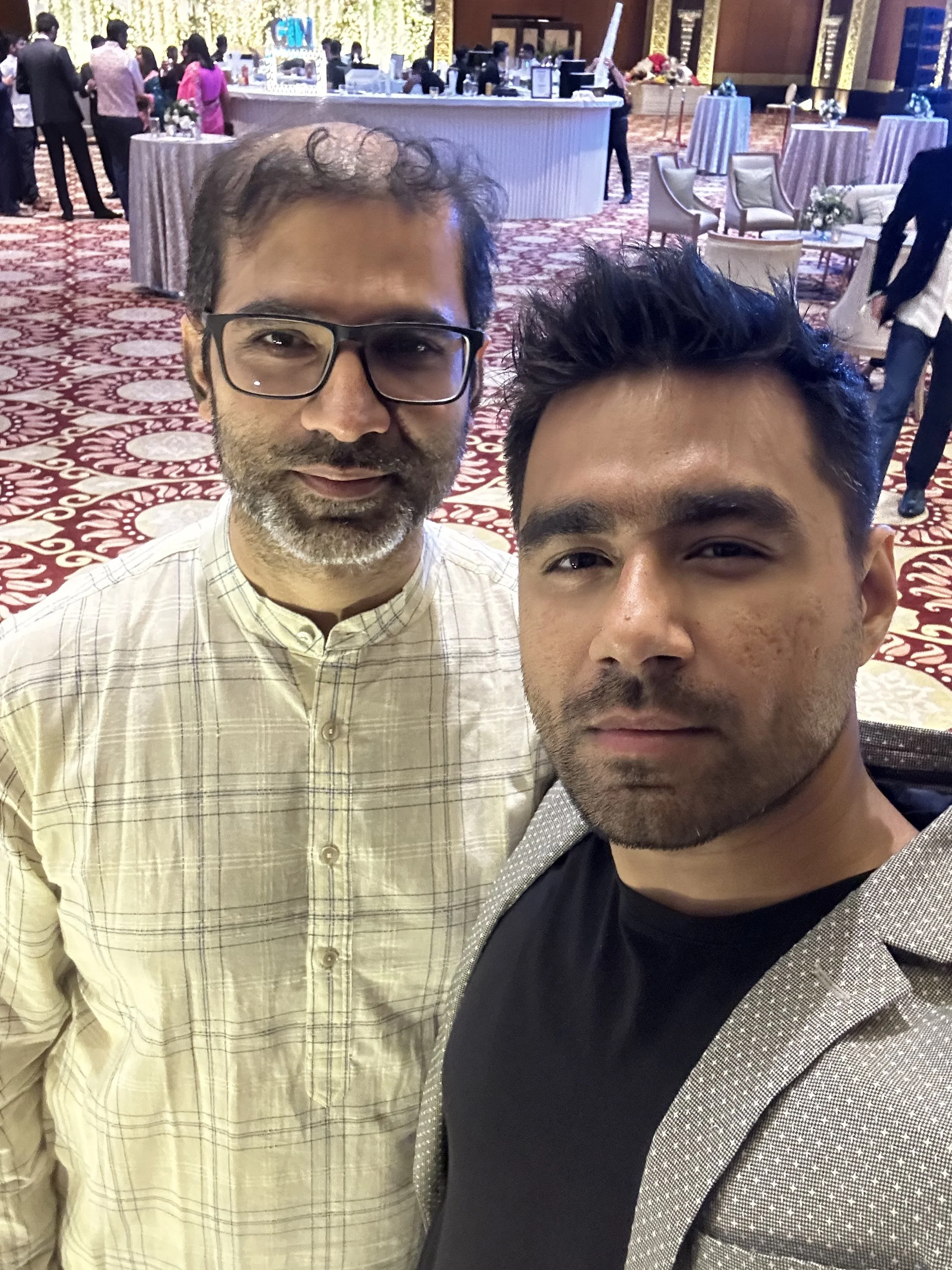 Two men taking a selfie in a large, well-lit event space with red and white patterned carpet, with many people, tables, and chairs in the background.